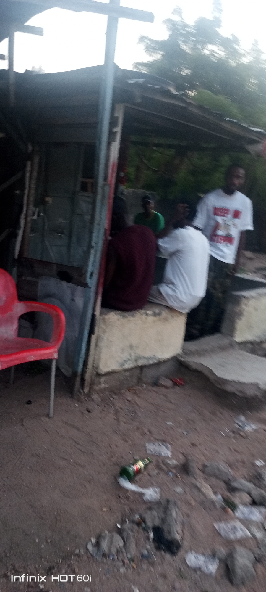 The image shows a makeshift roadside structure in Lokoja, Nigeria. The building appears to be constructed from corrugated metal and wood. Several people are present: one standing wearing a white t-shirt with a graphic print, two others sitting on a concrete ledge, and another person in a green shirt who is partially obscured. In front of the structure is a red plastic chair. Debris and litter, including a glass beer bottle, are scattered on the ground. The environment appears to be a dusty, unpaved area.