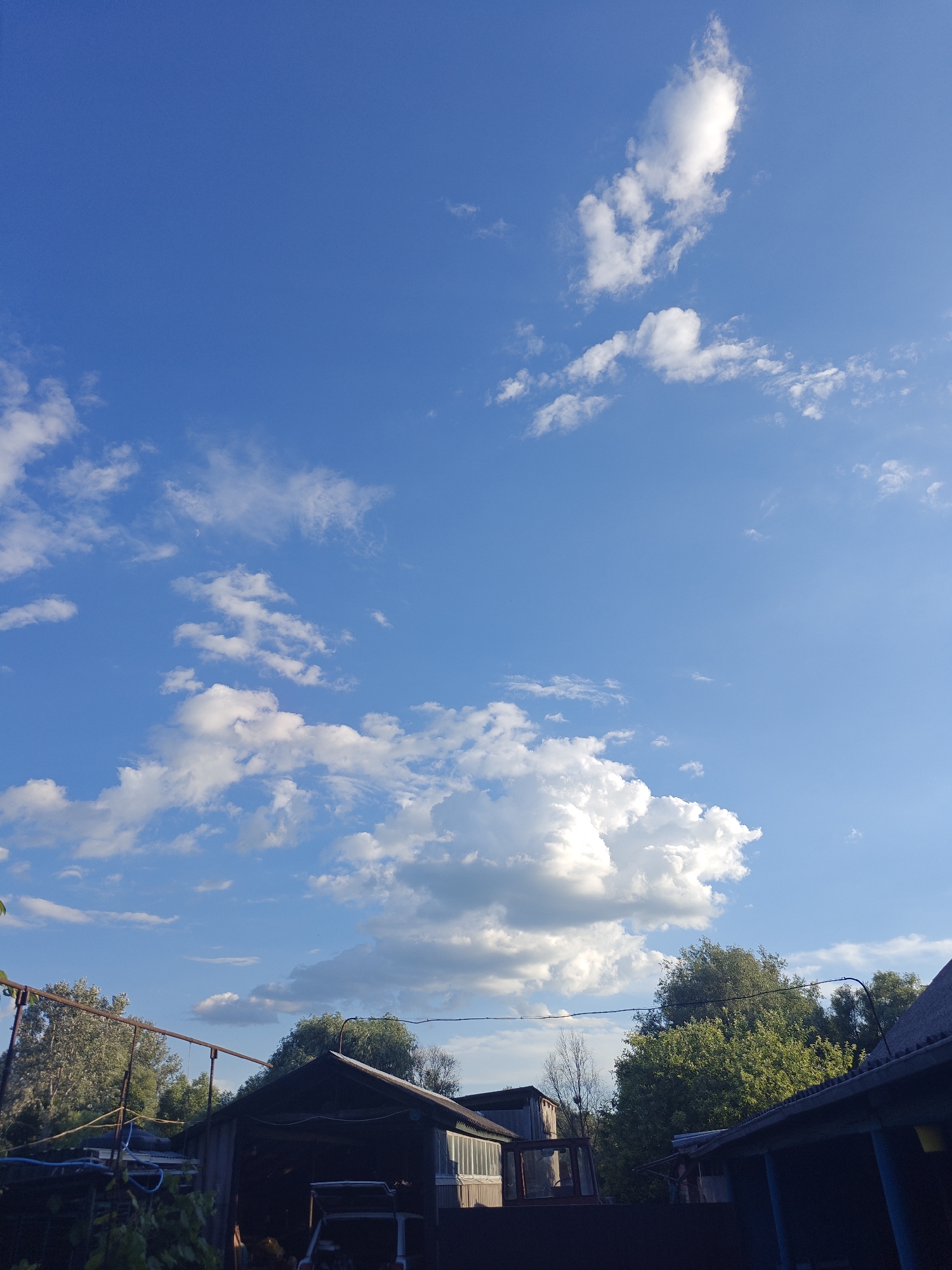 The image depicts a rural scene, likely a backyard in Kotelva, Ukraine, under a bright blue sky with scattered white clouds. In the foreground, there are several structures, including a dark-colored shed or garage with an open bay revealing items inside, and parts of another building with a corrugated roof. Trees, predominantly green deciduous ones, form a dense backdrop behind the structures, with some appearing drier and bare. A metal frame structure with horizontal bars is visible on the left side, possibly a clothesline or support. A small white vehicle, possibly a car or cart, is partially visible in the open bay of the shed. The lighting suggests it is daytime, with sunlight illuminating the clouds and casting shadows on the ground and structures. There are no people visible in the frame.