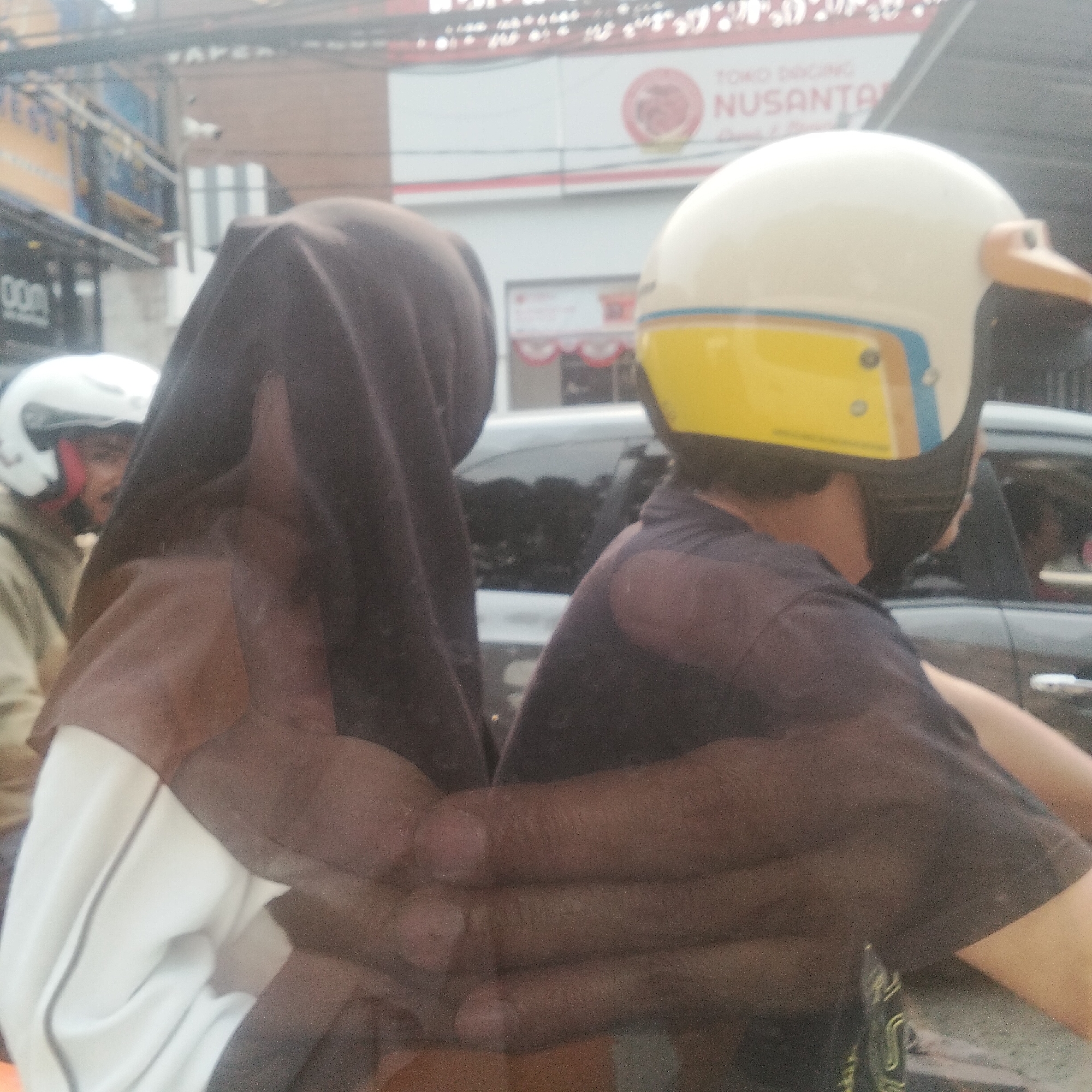 The image captures a reflection on a glass surface, likely from a vehicle, showing two individuals on a motorcycle. The foreground features the back of a person wearing a dark head covering and a white garment underneath. To the left, partially obscured, is another person wearing a white helmet with red trim and a light-colored jacket. In the background, a vehicle is visible, and beyond that, a street scene with buildings, signage, and streetlights. One sign displays "TOKO DAGING NUSANTARA" (Nusantara Meat Shop), a common type of establishment in Indonesian cities. The overall impression is that of a street scene in a city, likely Bogor, Indonesia, given the context of the establishment's name. The reflection creates a layered effect, making it challenging to discern the exact relationships or actions of the individuals, though they appear to be stationary on the motorcycle. The lighting suggests it is daytime, with a slight haze or glare obscuring some details.