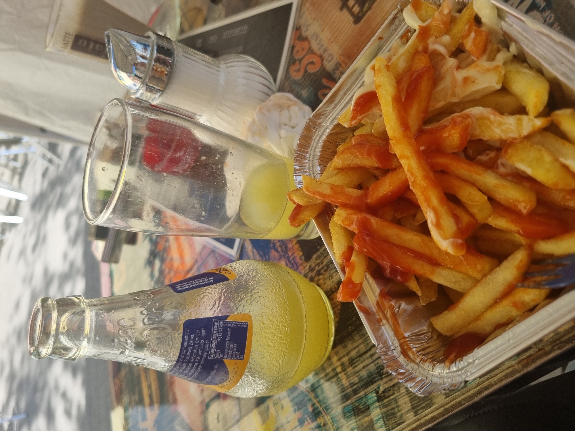 The image captures a casual outdoor meal scene, likely at a restaurant in Saarbrücken, Germany. An aluminum container filled with french fries, generously topped with ketchup and mayonnaise, sits on a patterned tabletop. A glass with a yellow liquid is partly filled. A bottle of Oranġina, a carbonated citrus beverage, lies on its side, its label visible with information in German. A sugar dispenser is also present. The composition of the shot is somewhat casual and likely taken from a diner's perspective. The environment appears to be a restaurant patio with the hint of a streetscape in the background.