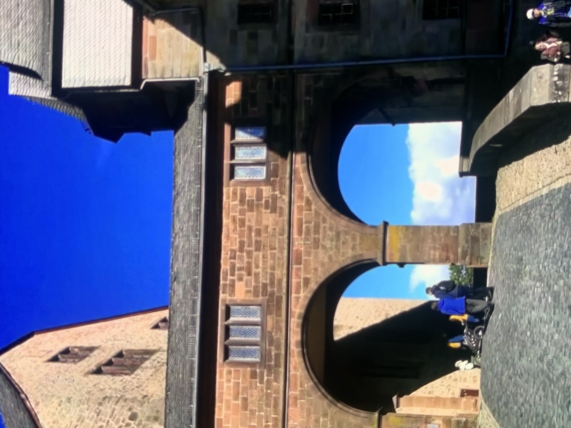 This image depicts a scenic view of a historic stone building with arched passageways against a bright blue sky. The architecture suggests a medieval or Renaissance style, with rough-hewn stone blocks and multi-paned windows. In the foreground, a cobblestone path leads up to the arches, where several people are gathered. One group, including a person in a blue jacket and another in a dark jacket, appears to be interacting near a stroller-like object. Further down the path, two individuals are visible, one in a pink scarf and the other wearing a baseball cap. The sky above is a vibrant, cloudless blue, offering a stark contrast to the earthy tones of the building. The overall atmosphere is one of historical grandeur and a pleasant day outdoors. This location is not in Stavropol, Russia.