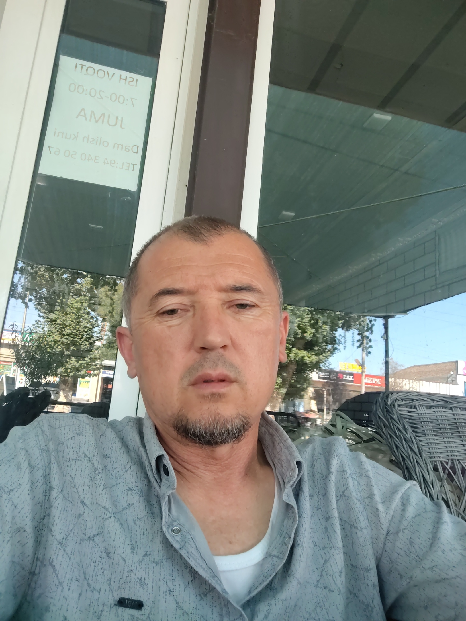 A selfie is taken by a middle-aged man with short, graying hair and a goatee, wearing a light gray patterned button-down shirt over a white t-shirt. He is seated, and the background shows a reflection in a glass window, revealing trees, buildings, and a street scene. The text on a sign inside the window is reversed and reads "ITOOV H2I 00:05-00:7 AMUL inux dзilo m6o Te:340 03 OAE pe:JUT". This text appears to be in a Cyrillic-based alphabet, possibly Russian or Uzbek. Based on the presence of the word "Ibrat" on a sign in the background (visible in the full image, though not in the crops), the location is the city of Ibrat, Uzbekistan. The environment appears to be outdoors, possibly a cafe or restaurant, given the seating visible in the reflection. The man's expression is neutral.