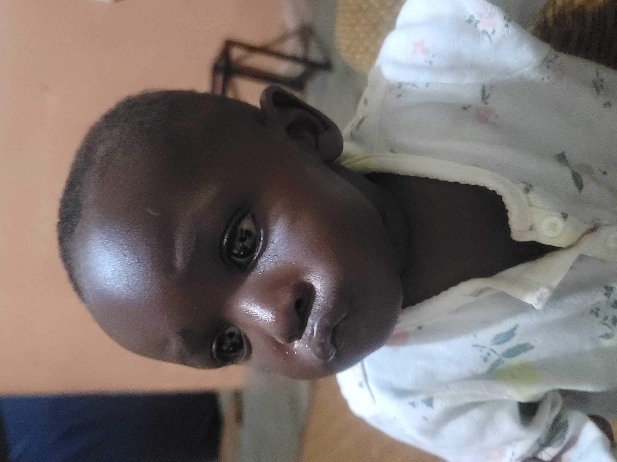The image shows a close-up, eye-level, medium shot of a young child's face, likely indoors in Barakin Akawo, Nigeria. The child has dark skin and appears to be around one year old. The child's lips are pursed, and their cheeks are slightly puffed out, giving them a somewhat pouting expression. There are visible droplets of moisture on the child's face, particularly around the nose and mouth, possibly indicating drool or a drink. The child is wearing a white onesie with a delicate floral pattern. The background is softly blurred, with a plain, peach-colored wall and a hint of a dark blue object on the left. A blurred wooden stool or chair with crisscrossed legs is visible in the mid-background. The lighting appears to be natural, possibly from a window, illuminating the child's face and creating a slight sheen on their skin.