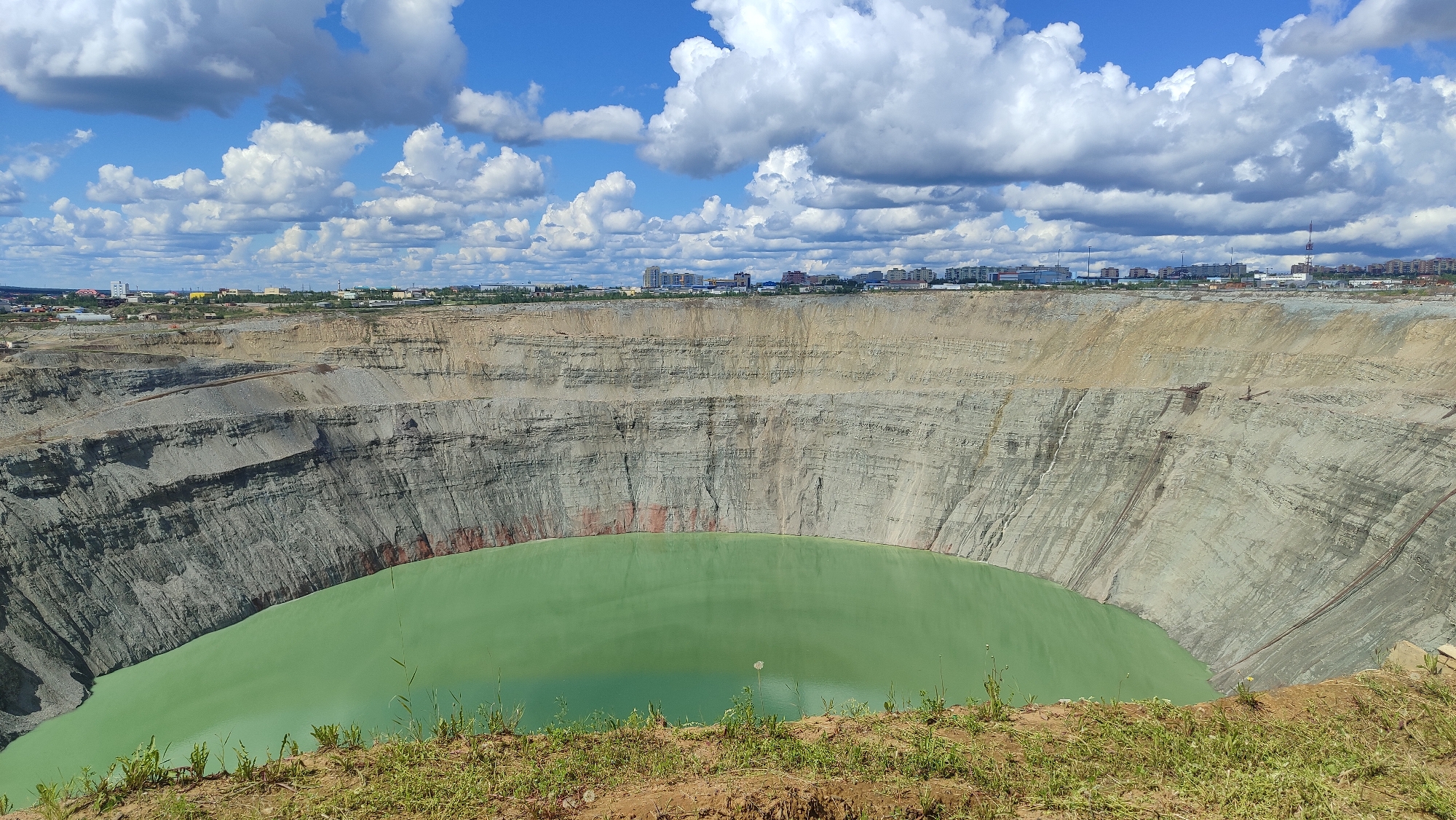 The image shows a vast, man-made open-pit mine, likely the Mir diamond mine, located in Mirny, Russia. The pit is circular with steep, layered rock walls that descend to a large body of murky, greenish water at the bottom. The scale of the excavation is evident from the distant city skyline visible on the horizon above the rim of the pit. The sky is blue with scattered white, puffy clouds. The foreground consists of rough, grassy terrain, suggesting the edge of the pit. No people are visible in the image. The scene depicts a significant industrial scar on the landscape, a testament to extensive mining operations.