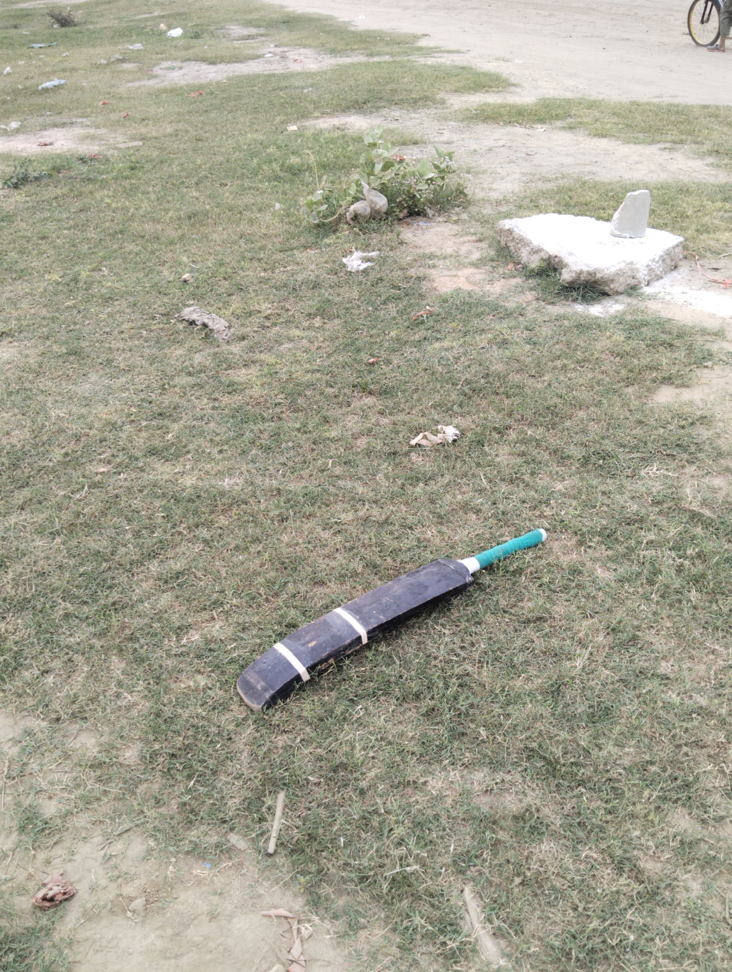The image shows a cricket bat lying on a grassy field. The bat is black with white stripes and a green handle. The field is uneven with patches of dry grass and dirt. A concrete structure is in the background, and the edge of a bicycle wheel is visible in the upper right corner. The location is Kot Duni Chand, Pakistan.