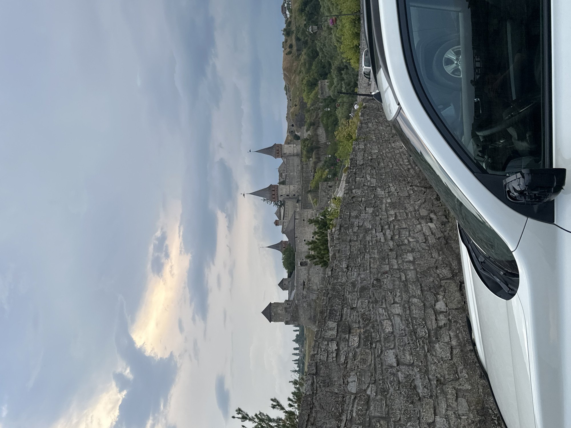 The image depicts a view of the Kamianets-Podilskyi Castle in Ukraine. A white SUV is partially visible in the foreground, with its roof rack and windshield wipers in focus. Behind the car, a stone wall runs horizontally across the frame. The castle is situated on a hill in the background, characterized by its stone towers and conical roofs, with a dramatic cloudy sky overhead. Green vegetation and trees surround the castle and the hilly landscape.