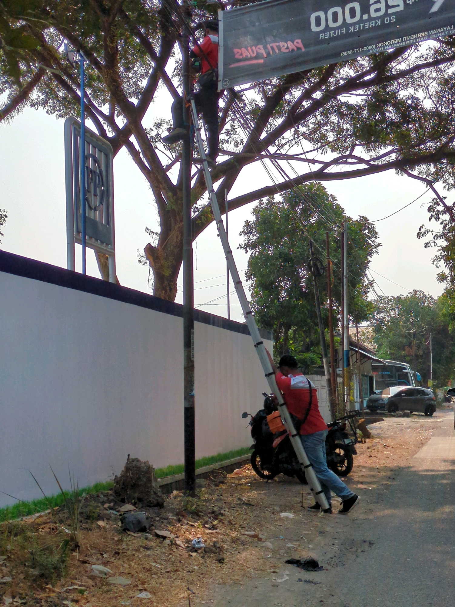 A person in a red shirt and blue jeans is carrying a long, silver ladder. They are walking on a dirt path next to a white wall and a street. A black motorcycle is parked nearby. In the background, there are trees, utility poles, and several cars and a bus. The image appears to be taken in Surakarta, Indonesia, given the style of the utility poles and the tropical foliage.