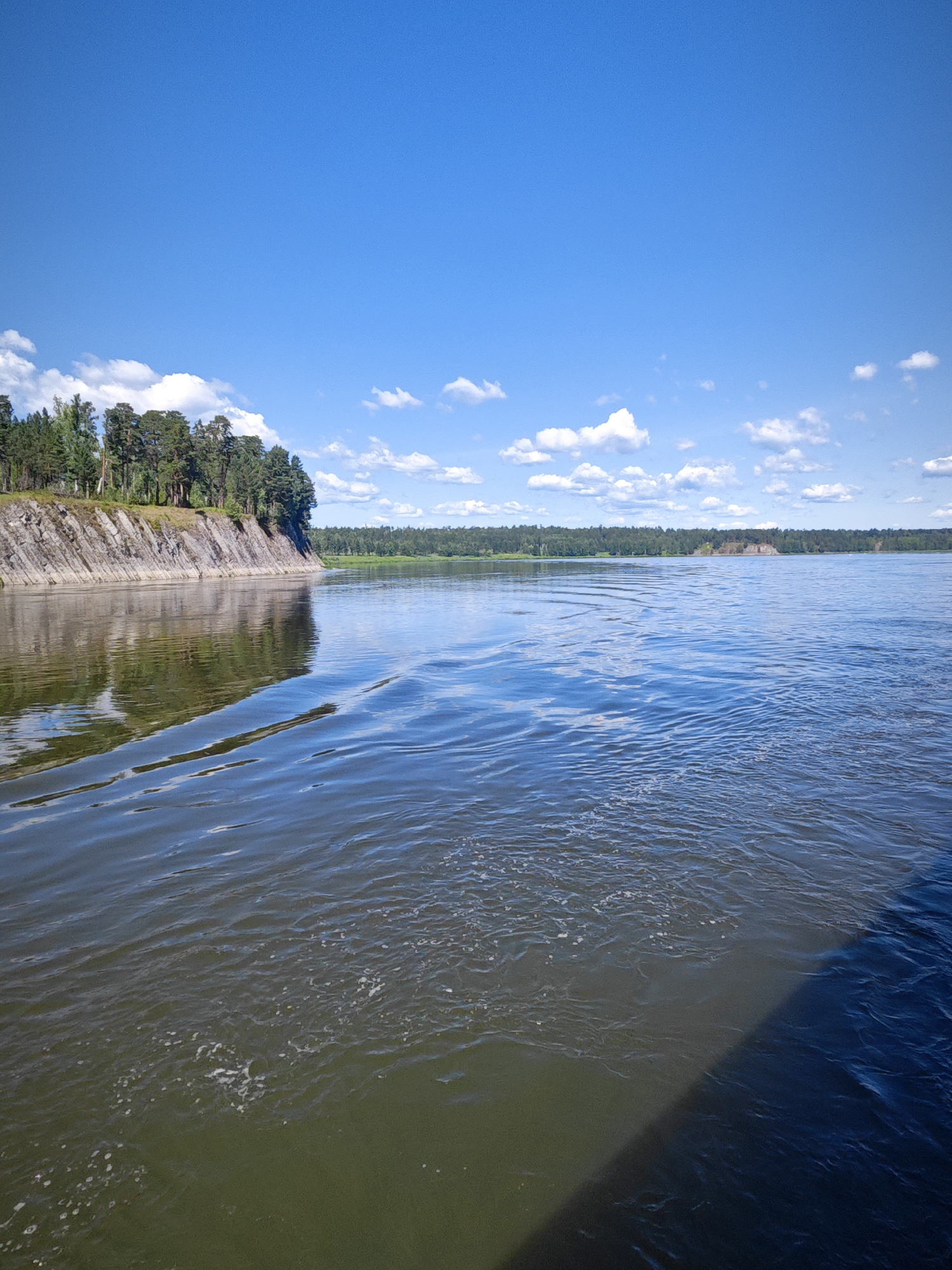 The image depicts a wide, calm river on a clear, sunny day. The sky is a vibrant blue with scattered white clouds. On the left bank, a steep, rocky cliff rises, topped with a dense forest of tall pine trees. The trees cast reflections on the surface of the water, which appears slightly murky and reflects the blue of the sky in its more distant sections. Ripples and small waves are visible on the water's surface, suggesting a gentle current or movement. The far bank of the river is also lined with a thick line of green trees. A diagonal shadow falls across the lower right portion of the water, likely cast by a boat or another unseen object. The scene appears to be a natural, undeveloped landscape. It is not possible to determine the specific location within the city of Savino, Russia from the visual information provided.