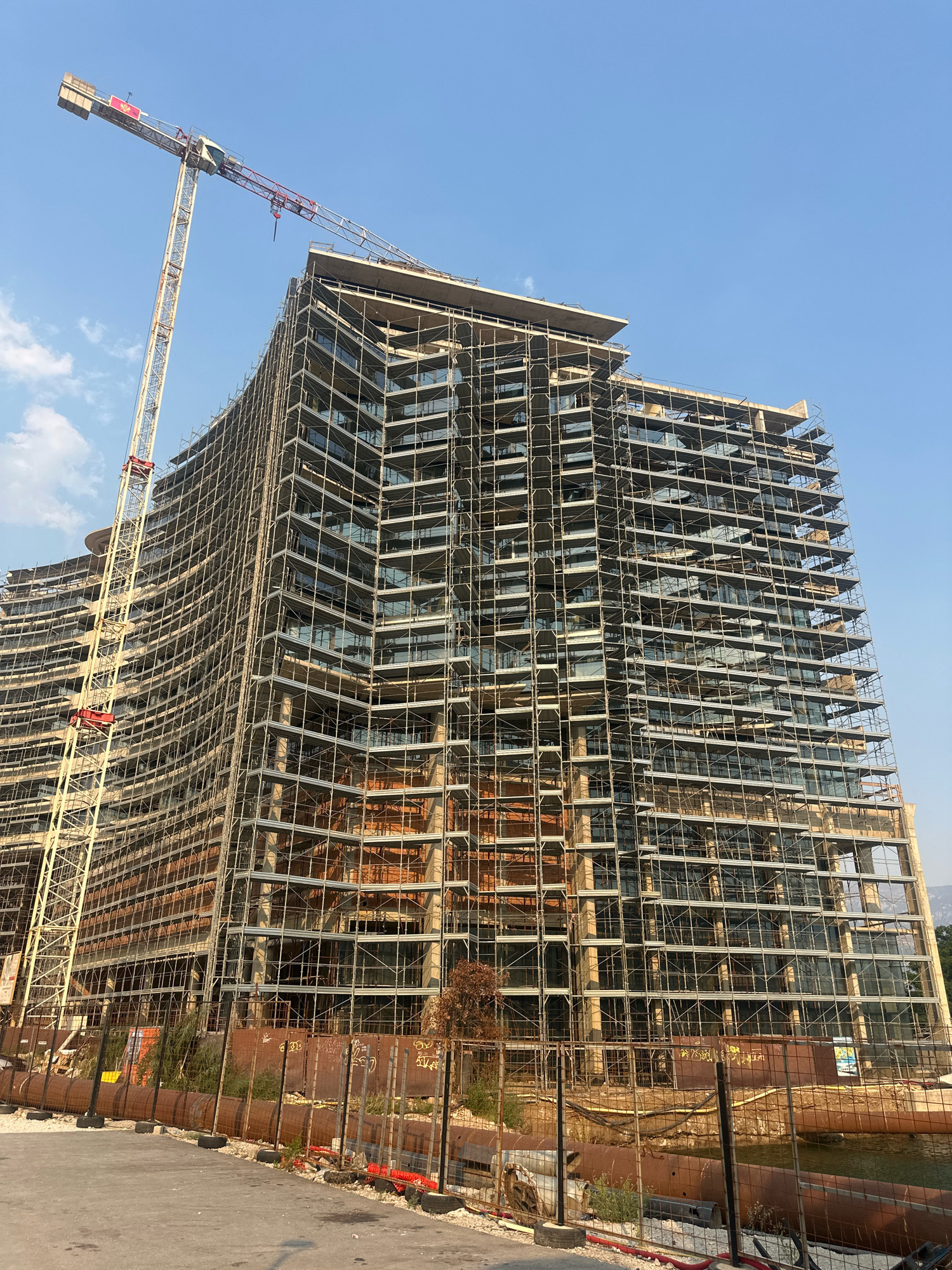 An exterior, low-angle shot captures a large building under construction in Budva, Montenegro. The building is encased in extensive scaffolding and appears to be multiple stories high. A tall construction crane is positioned to the left of the building, its jib extending over the structure. The sky is a clear, bright blue with a few wispy clouds visible. In the foreground, a fenced-off construction site is visible, with construction materials and debris present. The overall impression is of a busy construction site in an urban environment.