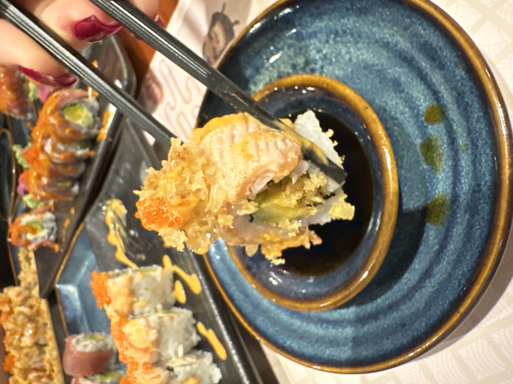 A close-up shot captures a hand with dark red nail polish holding black chopsticks, presenting a piece of sushi about to be dipped into soy sauce. The sushi roll features what appears to be a salmon topping over a layer of crispy, possibly tempura, flakes, with white rice and green filling (likely avocado or cucumber) visible underneath. The chopsticks precisely hold the sushi above a small, circular well of dark soy sauce, which is part of a larger, blue ceramic plate with a textured, spiral design and a distinct brown rim.
The background is softly blurred but shows an array of other sushi rolls arranged on dark, rectangular serving platters, suggesting a meal with multiple sushi varieties. Some rolls are topped with orange roe or drizzled with yellow sauces. The overall setting appears to be an indoor dining environment, likely a restaurant specializing in sushi or Japanese/Asian cuisine. The lighting is soft and ambient. No discernible text is visible. This scene depicts a dining experience in Nha Trang Ward, Vietnam.