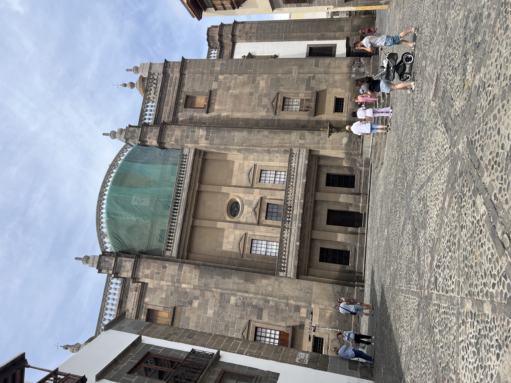This image depicts a large stone building in what appears to be the city of Las Palmas De Gran Canaria, Spain. The building, possibly a cathedral or church, features multiple windows, doors, and architectural details such as a dome currently under restoration covered by a green net.
Several people are visible in the image, including a person pushing a stroller, and several walking nearby. The ground is paved with uneven cobblestones. The sky is partly cloudy. The overall impression is of a historical building within a bustling public space.