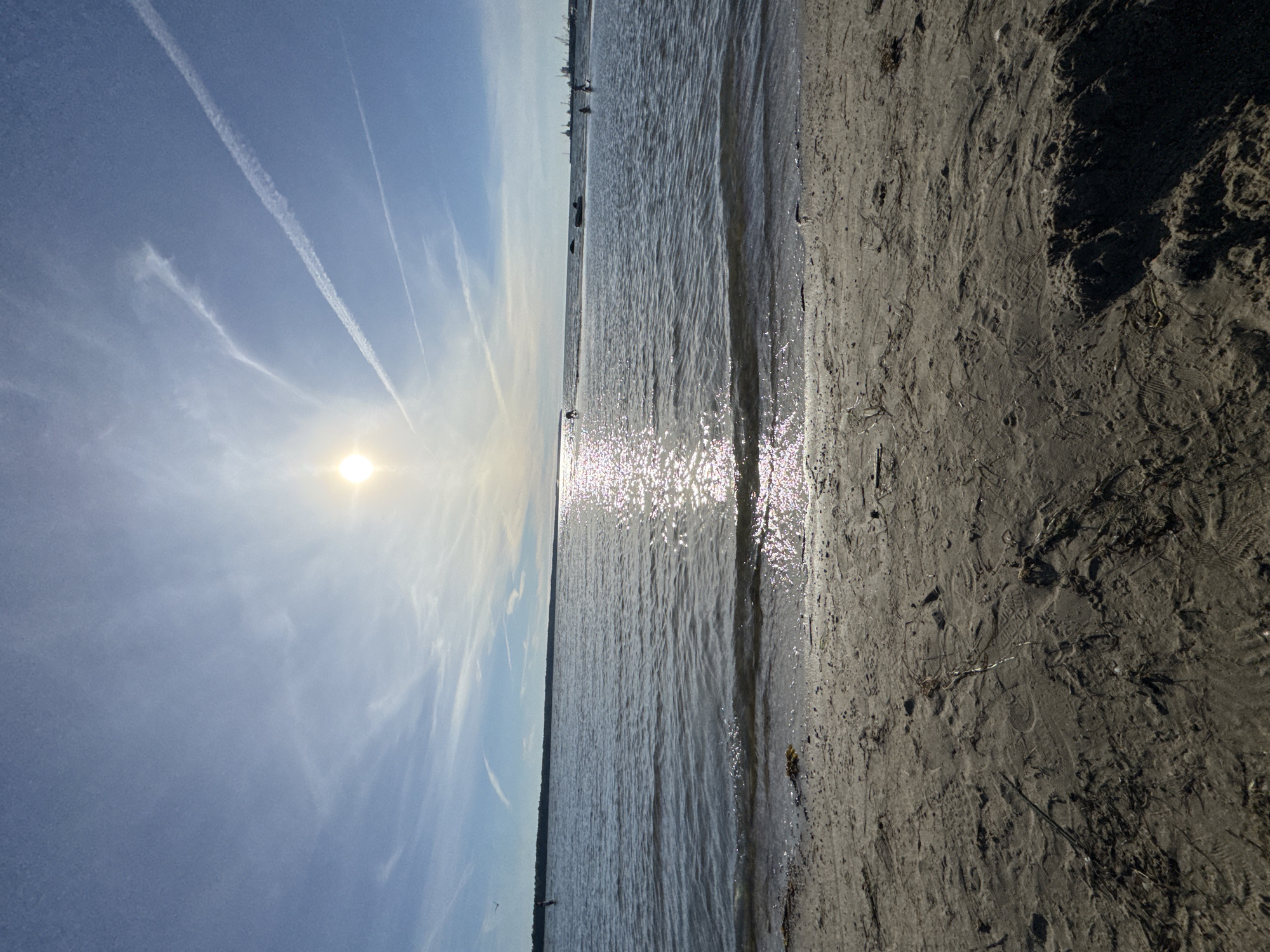 This is a landscape photograph taken on a sunny day at a beach, likely in Tallinn, Estonia, given the context of previous requests. The scene depicts a calm body of water, possibly the Baltic Sea, meeting a sandy shore in the foreground. The sun is bright and positioned high in the sky, creating a strong glare on the water's surface and casting shadows on the sand.
The sky is a clear blue with scattered wispy clouds and several prominent contrails from aircraft. The water is relatively shallow near the shore, with gentle waves lapping at the sand. In the distance, there are several people engaged in activities on or near the water. Some appear to be wading or playing, while others might be participating in water sports. Farther in the distance, there is a faint outline of a cityscape or industrial area with structures resembling cranes and buildings.
The sandy beach in the foreground is textured with footprints, debris, and uneven surfaces, indicating it's a public beach. The overall atmosphere is one of a bright, pleasant day at the seaside.