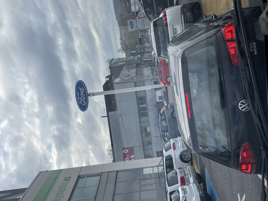 The image depicts an outdoor scene in a commercial district of Oiartzun, Spain, characterized by overcast weather. In the immediate foreground, the rear section of a dark grey or black Volkswagen Golf TDI is visible, including its illuminated red taillights and rear Volkswagen emblem. The perspective suggests the photograph was taken from within another vehicle.
Directly ahead of the Golf is a white Peugeot 3008 SUV, identifiable by its design and a partially visible Spanish license plate including "MKM". A queue of vehicles extends forward from this point, including a red hatchback and a blue sedan, among others. To the left, a large, multi-story building with grey paneling and numerous windows is visible, featuring a partial green sign displaying "oymerlin.es", indicating a Leroy Merlin store.
Central to the midground is a tall pole topped with a prominent blue oval "Ford" sign. Below this, another sign for "Ford Baso-Motor" is visible, alongside a "REPSOL" sign, indicating the presence of a Ford dealership and a fuel station. The background consists of distant, tree-covered hills under a uniformly grey, cloudy sky.