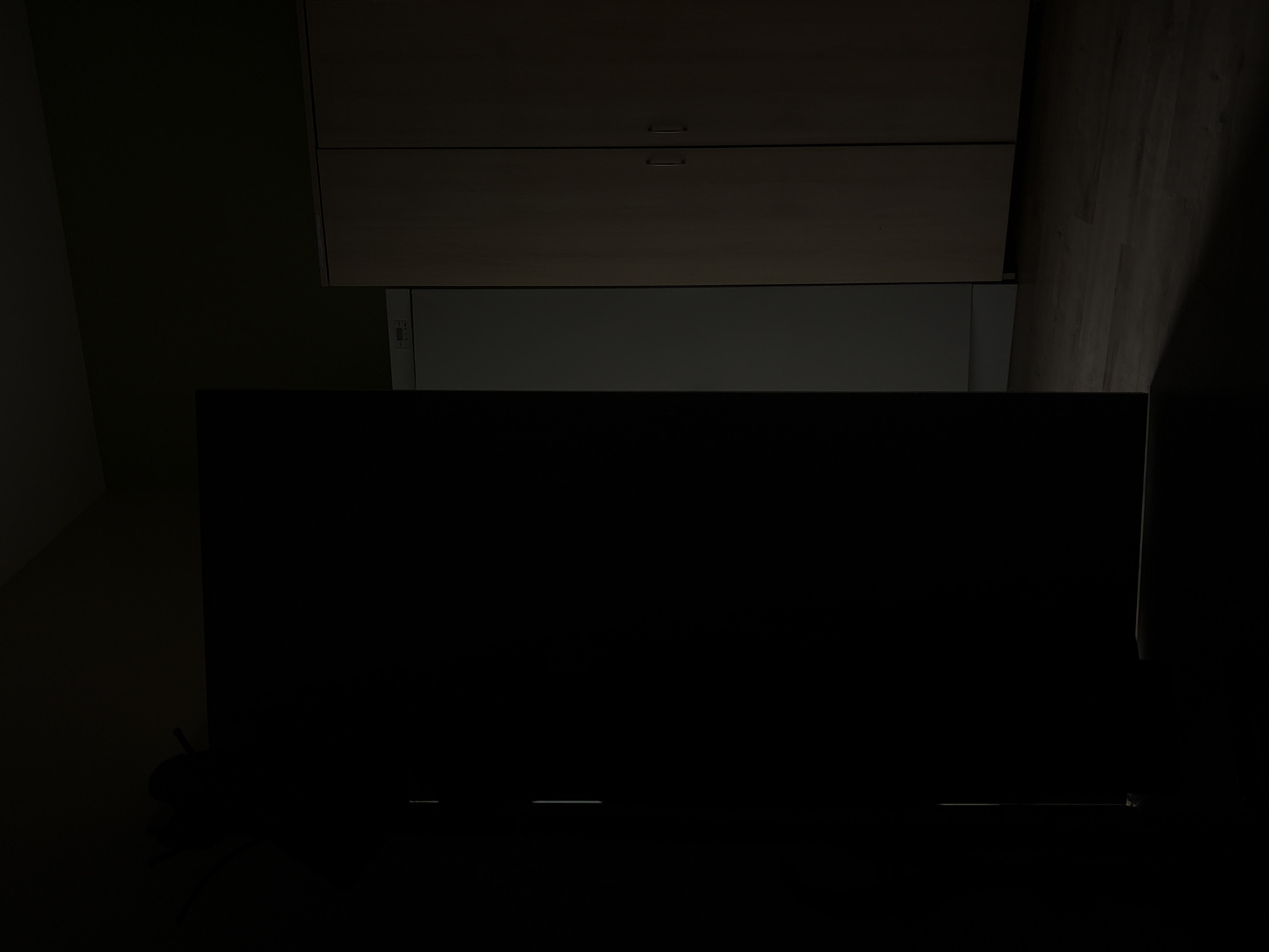 The image depicts an indoor scene, likely a room, characterized by low lighting. A large, dark, rectangular object, possibly a door or a panel, occupies the left side of the frame, casting a significant shadow. To the right of this object, a sliver of a white appliance, possibly a refrigerator or a washing machine, is visible. Adjacent to the appliance are two tall, light-colored cabinets or wardrobes, each with a single handle. The floor appears to be wooden planks. The overall impression is of a dimly lit interior space.
Regarding the location in the city of Aldan, Russia, there are no visual cues within the image that can be used to identify this specific location. The scene is generic and could be found in many residential or commercial interiors.