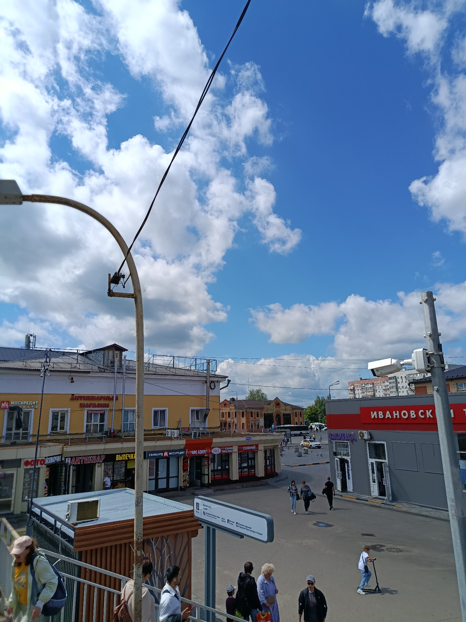 The image depicts a street scene in Orekhovo-Zuyevo, Russia. The sky is blue with scattered white clouds. In the foreground, a pedestrian walkway with metal railings is visible. Several people are walking on the walkway and in the street. On the street, there are buildings with various storefronts, including "МОСКРЕДИТ", "АНТИКВАРНЫЙ МАГАЗИН", "ФОТОКЛЮЧ", "ВЫПЕЧКА", and "СКЛАД". A sign with "ИВАНОВСКИ" is also visible on a building. A child is riding a scooter in the street. The scene appears to be a commercial area with pedestrian and vehicular traffic.
