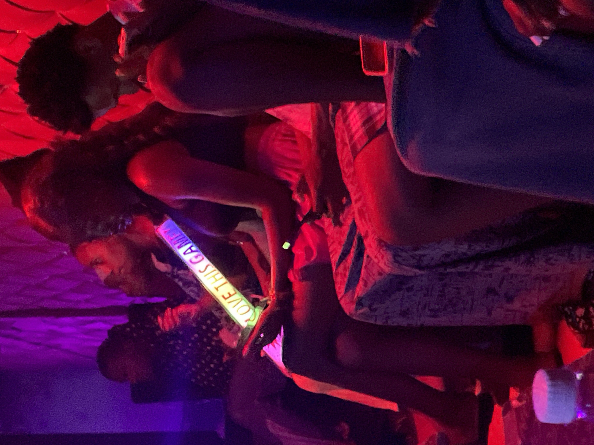 In Durumi, Nigeria, a group of people are gathered in a dimly lit environment with vibrant pink and red lighting. A woman in the foreground holds a neon glow stick that reads "LOVE THIS GAME" in capital letters. She is seated next to another individual, whose legs are visible and wearing checkered shorts. To the right, a man with curly hair is partially visible, looking down. In the background, other individuals are seated, with one person wearing a patterned shirt. The overall atmosphere suggests a social gathering or event taking place in a club or entertainment venue.