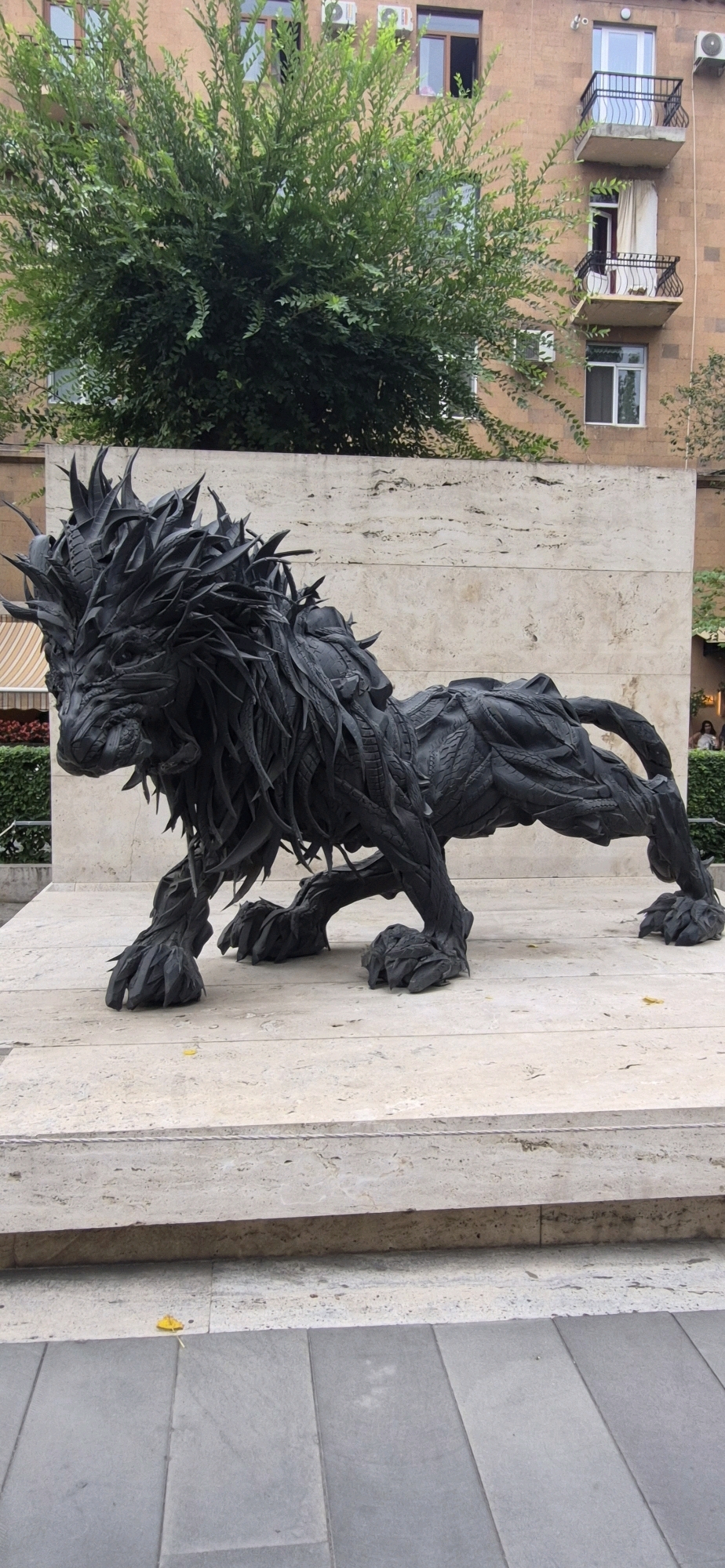 The image displays a large, black sculpture of a lion in a crouched position, made from what appears to be recycled tires. The sculpture is placed on a light-colored stone pedestal. Behind the sculpture, there is a beige stone wall and a lush green tree. In the background, a multi-story building with balconies and windows is visible. The ground in front of the pedestal is paved with grey rectangular tiles. This sculpture is located in Yerevan, Armenia, and is a well-known landmark. There are no people or animals clearly visible in the frame.