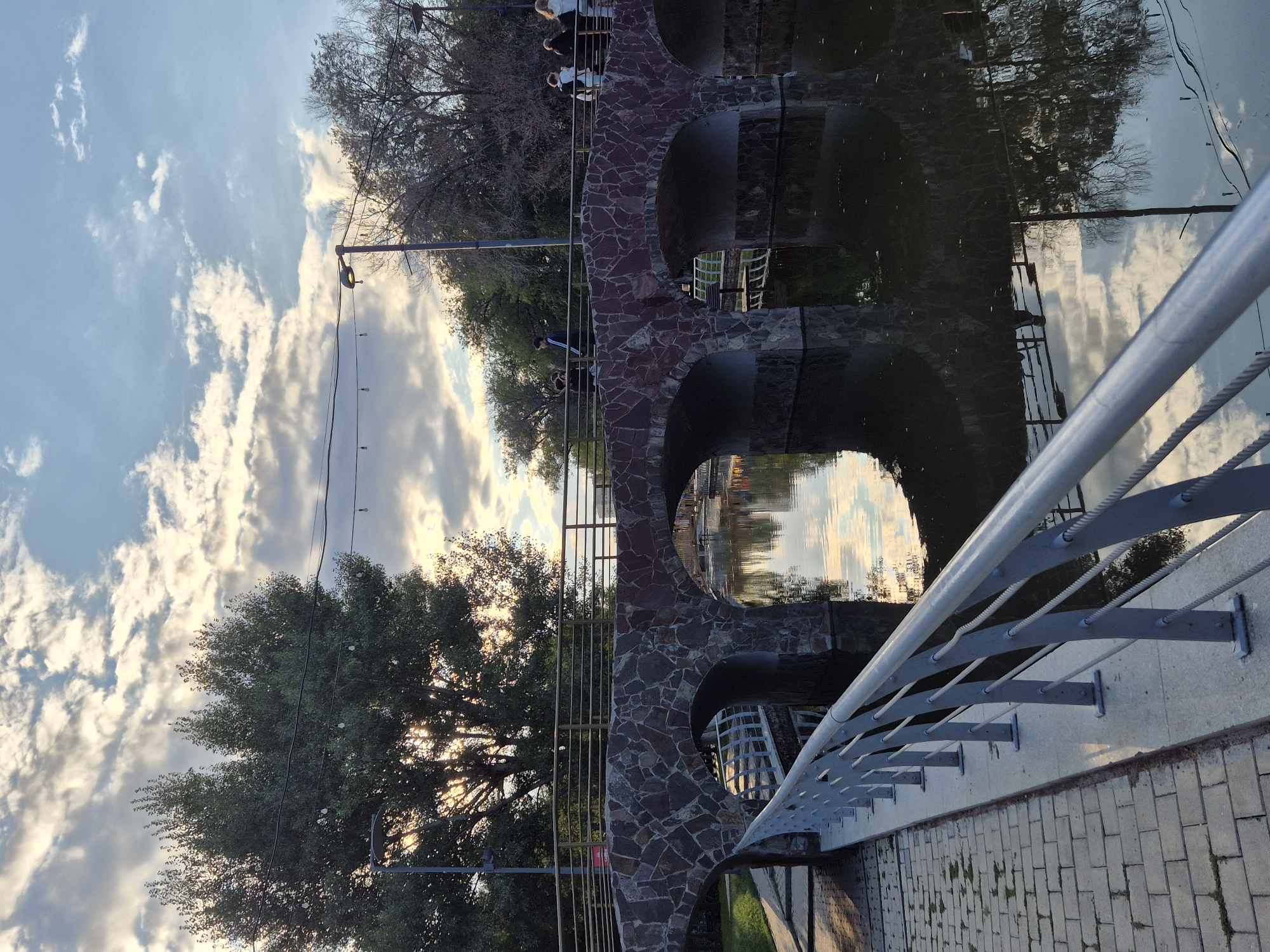 The image captures a view of a stone arch bridge spanning a body of water, likely a canal or river, in Astana, Kazakhstan. The bridge is constructed with rough-hewn, reddish-brown stones and features multiple arched openings. A metal railing runs along the top of the bridge, and several individuals are walking across it.
In the foreground, a grey metal railing lines a paved walkway, which appears to be made of light-colored rectangular stones. The water below reflects the bridge, the surrounding trees, and the sky. The sky is partially cloudy with patches of blue, and the sun is setting or rising, casting a warm glow and creating dappled light through the trees on the left.
The trees on the left side of the image are large and full of green leaves, while trees on the right appear more sparse, with some bare branches. Several streetlights and overhead wires are visible against the sky. On the bridge, there are at least four people visible, walking in different directions. One person on the far left is wearing dark clothing with a white stripe. To their right, a person in a black jacket and white trousers is visible. Further to the right, two individuals in white jackets are walking, and another person in dark clothing is seen behind them.
The overall scene suggests a public park or promenade area within the city, offering a tranquil atmosphere with the interplay of natural elements and man-made structures.