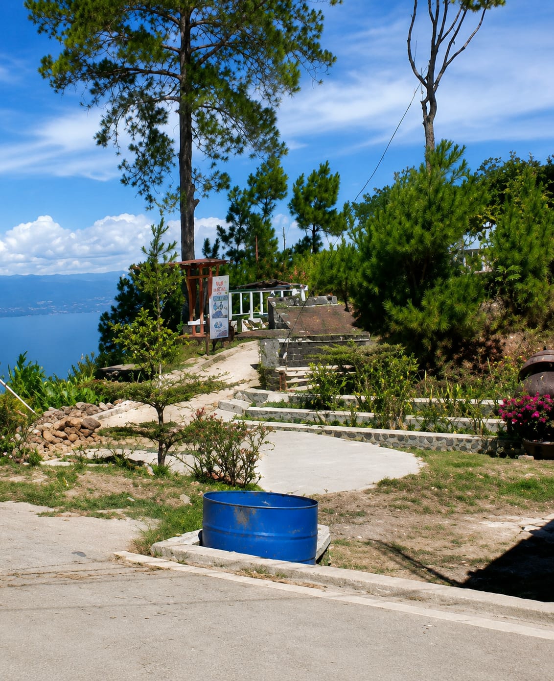The image depicts an outdoor scene in Parik Sabungan, Indonesia, featuring a terraced landscape with a large tree in the foreground. The terrain is covered with sparse grass, dirt, and some gravel. In the mid-ground, a winding concrete path leads up to a slightly elevated area with stone retaining walls and steps. Several bushes and small trees are planted along the path and terraces. A prominent blue cylindrical object, resembling a large barrel, sits on a concrete base near the bottom of the image. In the background, a body of water, likely a lake, is visible, bordered by distant hills. The sky is blue with scattered white clouds. The overall impression is of a scenic overlook or park area.