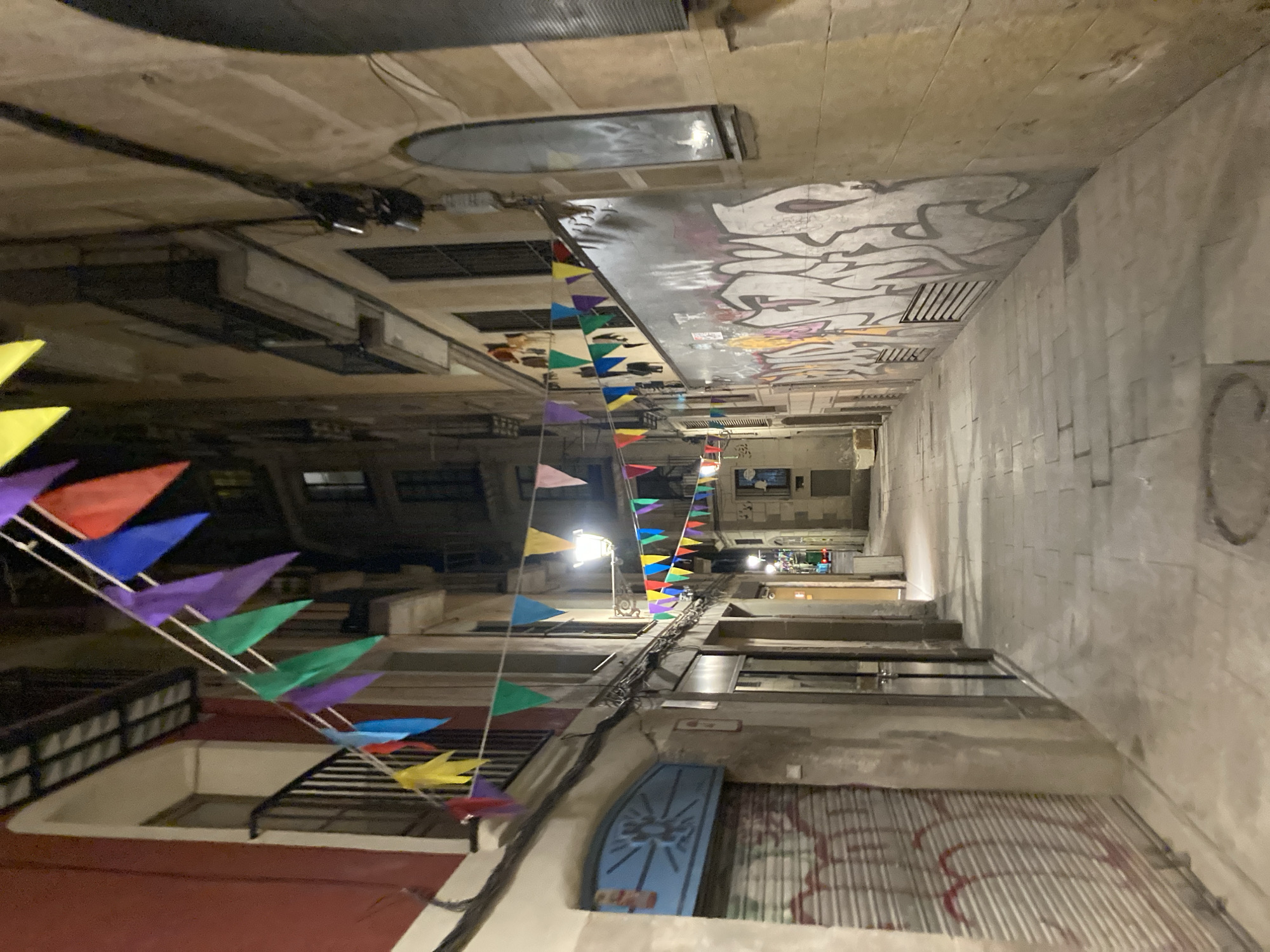 This image depicts a narrow, cobblestone alleyway in Barcelona, Spain at night. Festive, multicolored triangular flags are strung across the alleyway, adding a festive atmosphere. The buildings on either side of the alley are tall and have multiple stories with balconies. One building on the left has a red facade and a blue, sunburst-like design over a roll-down metal shutter. The building on the right features a large graffiti mural with abstract shapes and bold lines. Streetlights cast an artificial glow, illuminating the scene and creating shadows. A lone streetlamp is visible further down the alley, and the road ahead appears to be lit by more distant lights. The alleyway appears to be devoid of people.