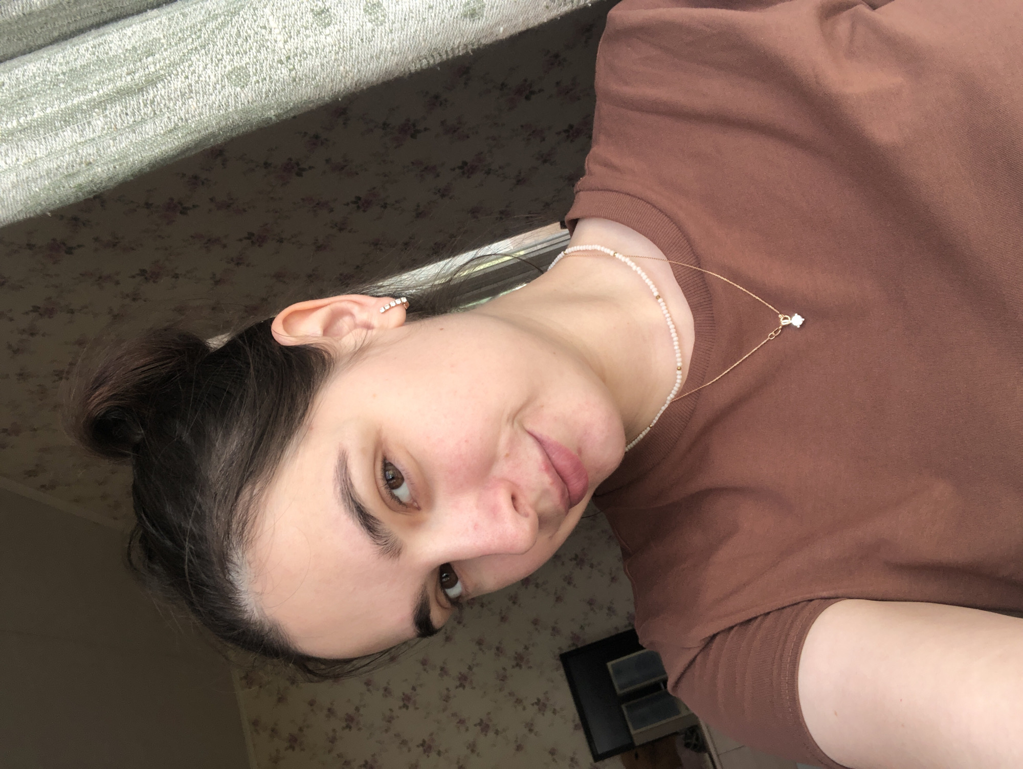 A young woman is captured in a close-up selfie, looking directly at the camera with a slight, neutral expression, suggesting a subtle smirk. Her head is tilted, indicating she is likely holding the camera herself. She has dark brown hair, styled in a bun or ponytail, with a few strands framing her face. Her eyebrows are dark and well-defined, complementing her brown eyes. Her complexion shows a natural flush on her cheeks and nose.
She is wearing a plain, short-sleeved brown t-shirt. Two necklaces adorn her neck: one is a delicate string of small, light-colored beads, and the other is a thin gold chain with a small, sparkling pendant, possibly a diamond or crystal. A small, multi-stone earring is visible in her left ear.
The setting appears to be indoors in Noviy, Russia. The background features a dark floral wallpaper or fabric with lighter floral motifs. A textured, light-colored fabric, possibly a curtain or window dressing, runs across the top of the image. A noticeable shadow falls across the floral pattern, suggesting natural light is entering from a window. In the lower right corner, a dark, rectangular object, possibly furniture or electronics, is partially visible. There is no visible text in the image.