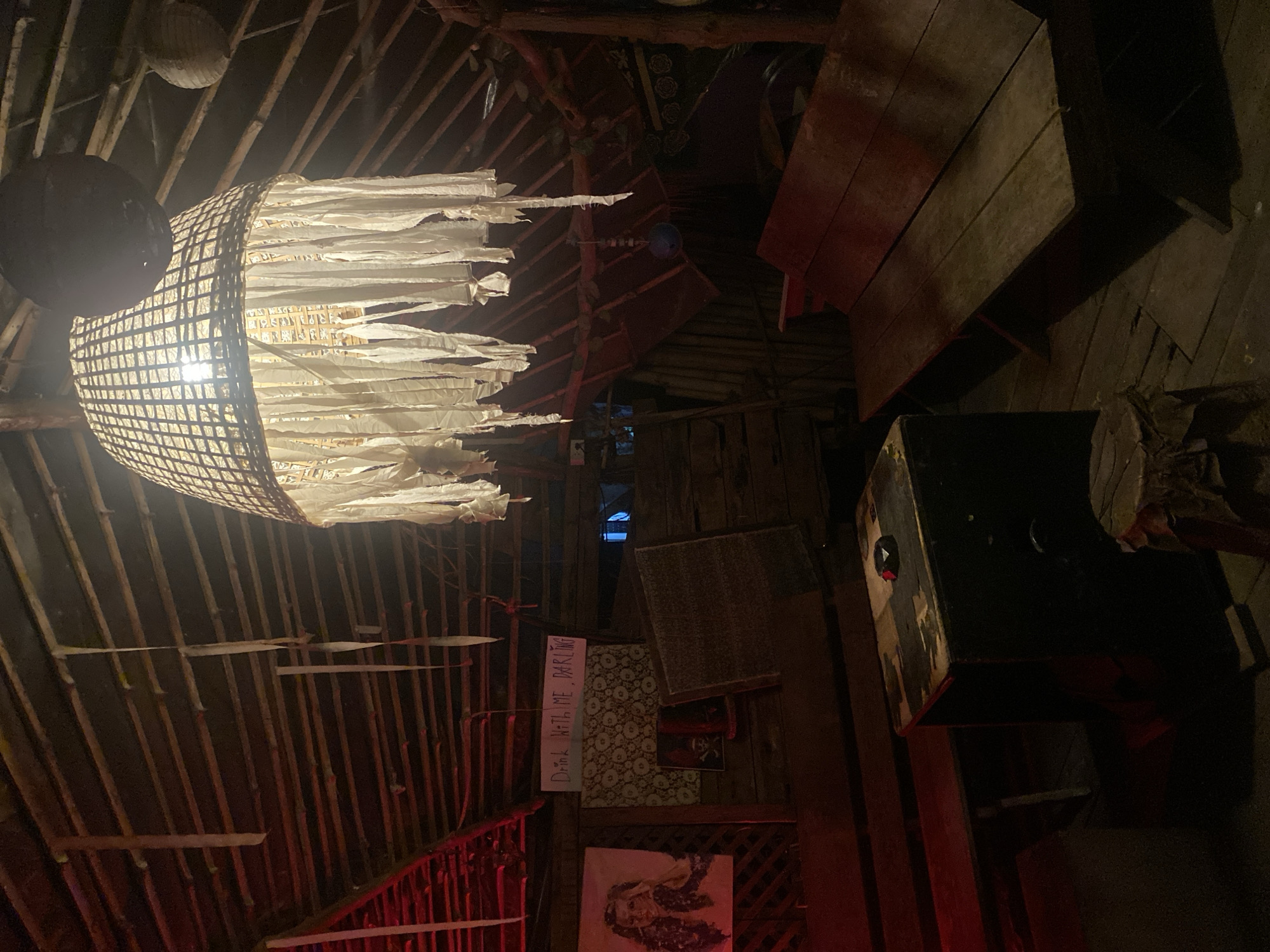 The image depicts the interior of a building in Baan Don Sai, Thailand. The structure appears to be constructed primarily of wood and bamboo. A unique chandelier-like light fixture hangs from the ceiling, with a woven shade and tattered white fabric strips.
To the right, a wooden bench or table is visible along with a dark box-like object. Further back, on the wall, are a sign with the words "Drink with me, darling", a patterned wallpaper, and framed images. The environment is dimly lit with a rustic, possibly bohemian, aesthetic.