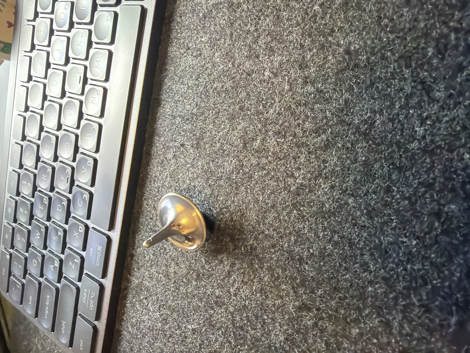 The image displays a close-up, angled view of a workspace, likely a desk, featuring a dark-colored keyboard and a metallic spinning top resting on a textured, grey surface.
The primary subject on the left is a portion of a keyboard, oriented vertically. It has dark grey keys with white lettering. The keyboard layout is QWERTY with secondary Hebrew characters, such as 'ש' (Shin), 'ד' (Dalet), 'ג' (Gimel), 'כ' (Kaf), 'ע' (Ayin), 'ח' (Chet), 'י' (Yod), 'ל' (Lamed), 'ם' (Mem Sofit), 'פ' (Pei), 'ף' (Pei Sofit), 'ת' (Tav), 'צ' (Tsadi), 'מ' (Mem), 'נ' (Nun), 'ב' (Bet), 'ס' (Samekh), 'ז' (Zayin), 'ק' (Kof), 'ר' (Resh), 'א' (Aleph), 'ט' (Tet), 'ו' (Vav), 'ן' (Nun Sofit). Visible English function keys include "tab," "caps lock," "shift," "ctrl," "start," "opt," "alt," and "cmd".
To the right of the keyboard, positioned centrally in the frame, is a small, silver-toned metallic spinning top, or a dreidel-like object. It has a shiny, reflective surface, indicating a polished metal construction, and is resting on its side with its pointed stem facing upwards and slightly left.
The surface beneath the keyboard and spinning top is a dark grey, highly textured material resembling a dense, short-pile carpet or a fabric desk mat. The lighting appears to be even, likely indoor, casting soft reflections on the metallic top. The presence of Hebrew characters on the keyboard strongly suggests the location is in Israel, aligning with the provided context of Kafr Yasif.