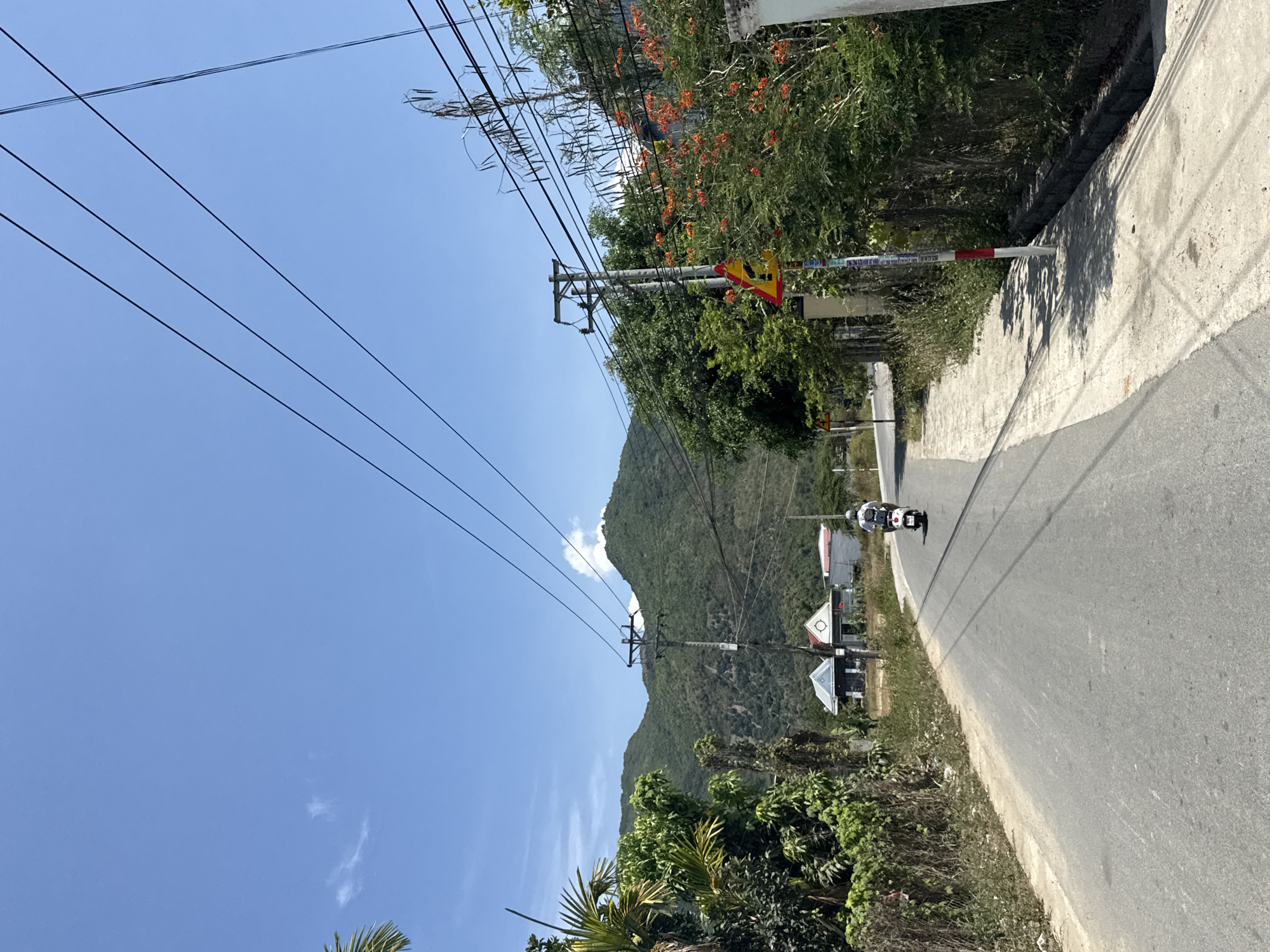 The image depicts a street in Diên Khánh, Vietnam, featuring a two-lane asphalt road curving to the right. A motor scooter with two people is traveling along the road. The road is bordered on the right by a concrete wall and vegetation, and on the left by trees and utility poles with power lines extending across the blue sky. A mountain is visible in the background. A road sign is present.