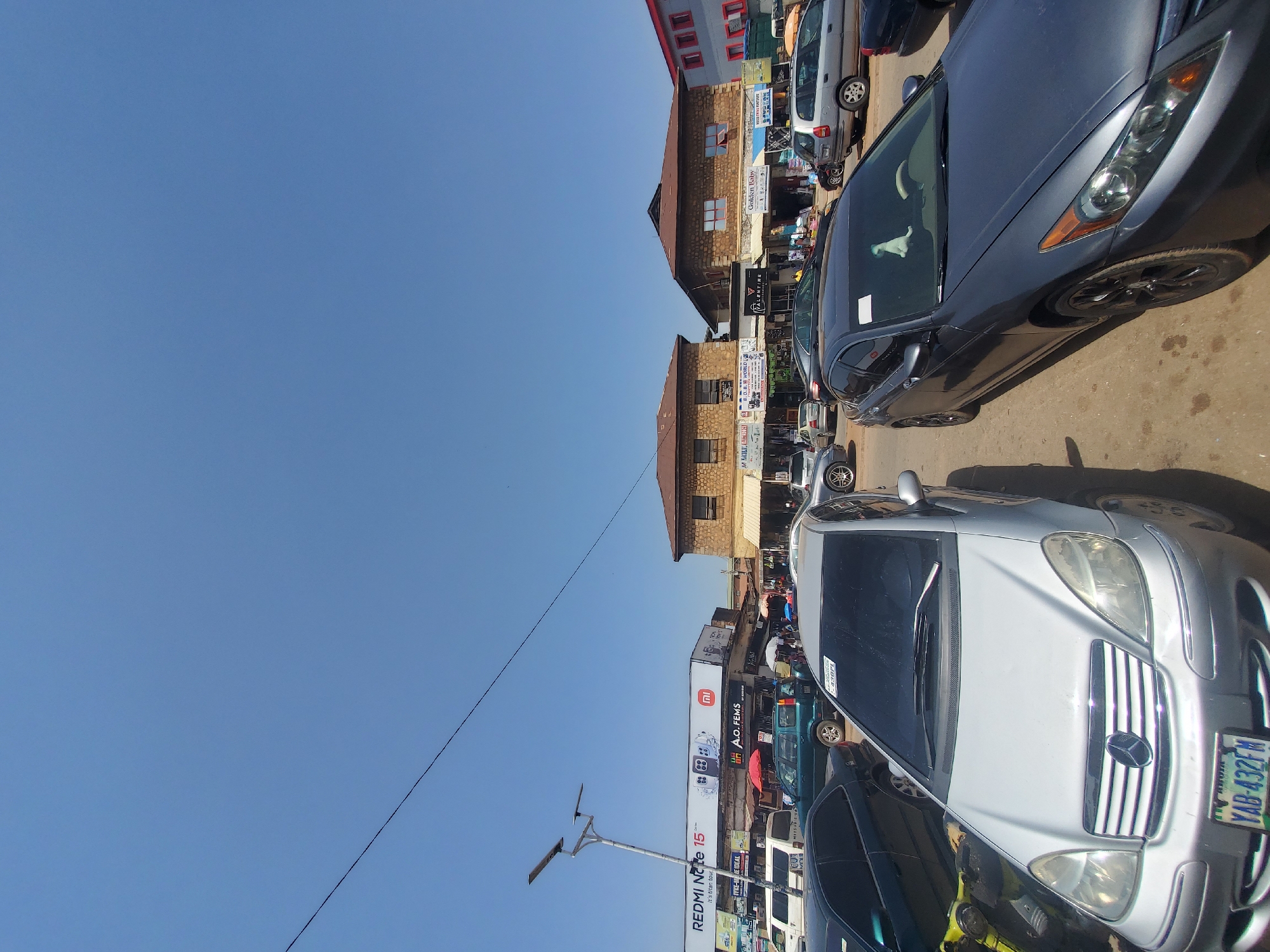 The image depicts a street scene in the city of Jos, Nigeria, under a clear blue sky. The foreground is dominated by several parked cars, including a silver Mercedes-Benz with license plate YAB-432FM and a dark gray sedan. In the background, a row of buildings lines the street, housing various businesses. Signs visible include "REDMI Note 15", "A.O. FEMS", "VALENTINE", and "GOLDEN BABY". The street appears to be a commercial area with vendors and shoppers present, indicated by the presence of umbrellas and people milling around. Power lines are visible crisscrossing the sky. The overall impression is that of a bustling marketplace in Jos.
