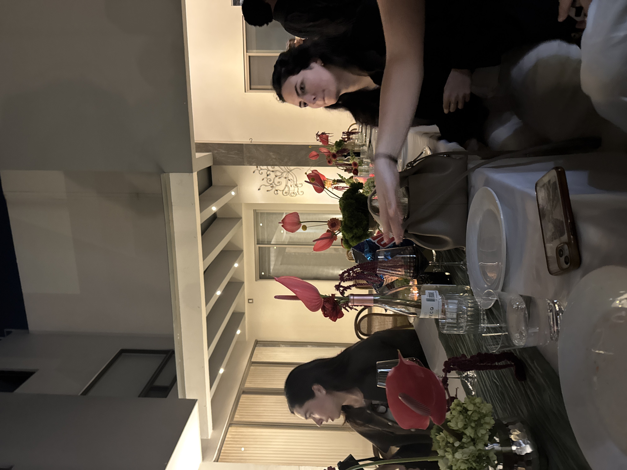 The scene depicts an evening social gathering around a dining table. The precise location within the city of Puebla, Mexico, is not visually discernible.
Three women are visible at the table. On the left, a woman with long, dark hair, wearing a dark top, is seen in profile, looking towards the left of the frame. On the right, a woman with long, dark, wavy hair, also in a dark top, is looking towards the right, her mouth slightly open. In the immediate foreground on the right, an arm and hand, belonging to an unseen person, extend across the table with the palm facing down. Partially visible in the background right is a person with dark hair.
The table is covered with a white tablecloth and a dark green, textured table runner. Multiple centerpieces feature red anthurium flowers, some accompanied by green floral clusters. Items on the table include a bottle of "MECQ" wine, several wine glasses (some containing red wine), water glasses, and empty white dinner plates. A small, dark red, human-like figurine stands on the table runner. A light brown handbag with a shoulder strap rests on the table near the woman on the right. A smartphone in a clear case is positioned on the table in the foreground.
The environment is an indoor or semi-outdoor space characterized by modern architecture. Light-colored walls, large windows or sliding doors, and a slatted ceiling with recessed lighting are visible. A decorative metal scrollwork piece is affixed to a wall in the background. The artificial lighting indicates a nighttime setting.
