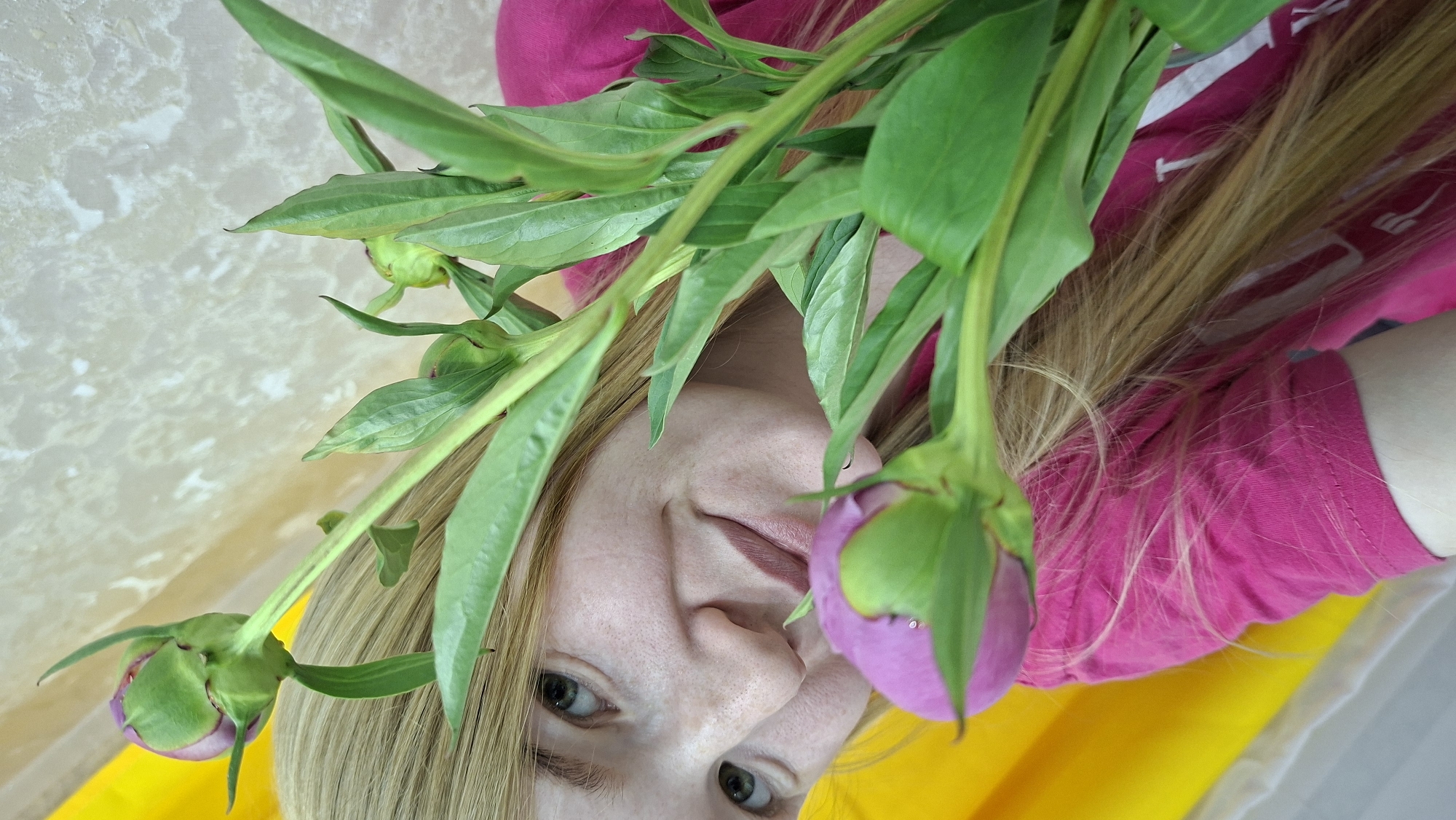 A fair-skinned female subject with long blonde hair and green eyes is depicted in a close-up, slightly tilted portrait. She wears a fuchsia-pink short-sleeved top. Her face, showing visible pores and a subtle smile with light-colored lips, is partially obscured by a bouquet of green-stemmed flowers. Three primary peony buds are visible, two with closed purple/pink petals encased in green sepals, and one smaller green bud further back. The subject's left eye, nose, and mouth are clearly visible, while her right eye is partially hidden by green foliage. To the immediate left of the subject, a bright yellow fabric or curtain is visible. The background consists of a light beige or off-white wall with a textured pattern. The specific location in the city of Chaikovsky, Russia, is not discernible from the visual information provided.