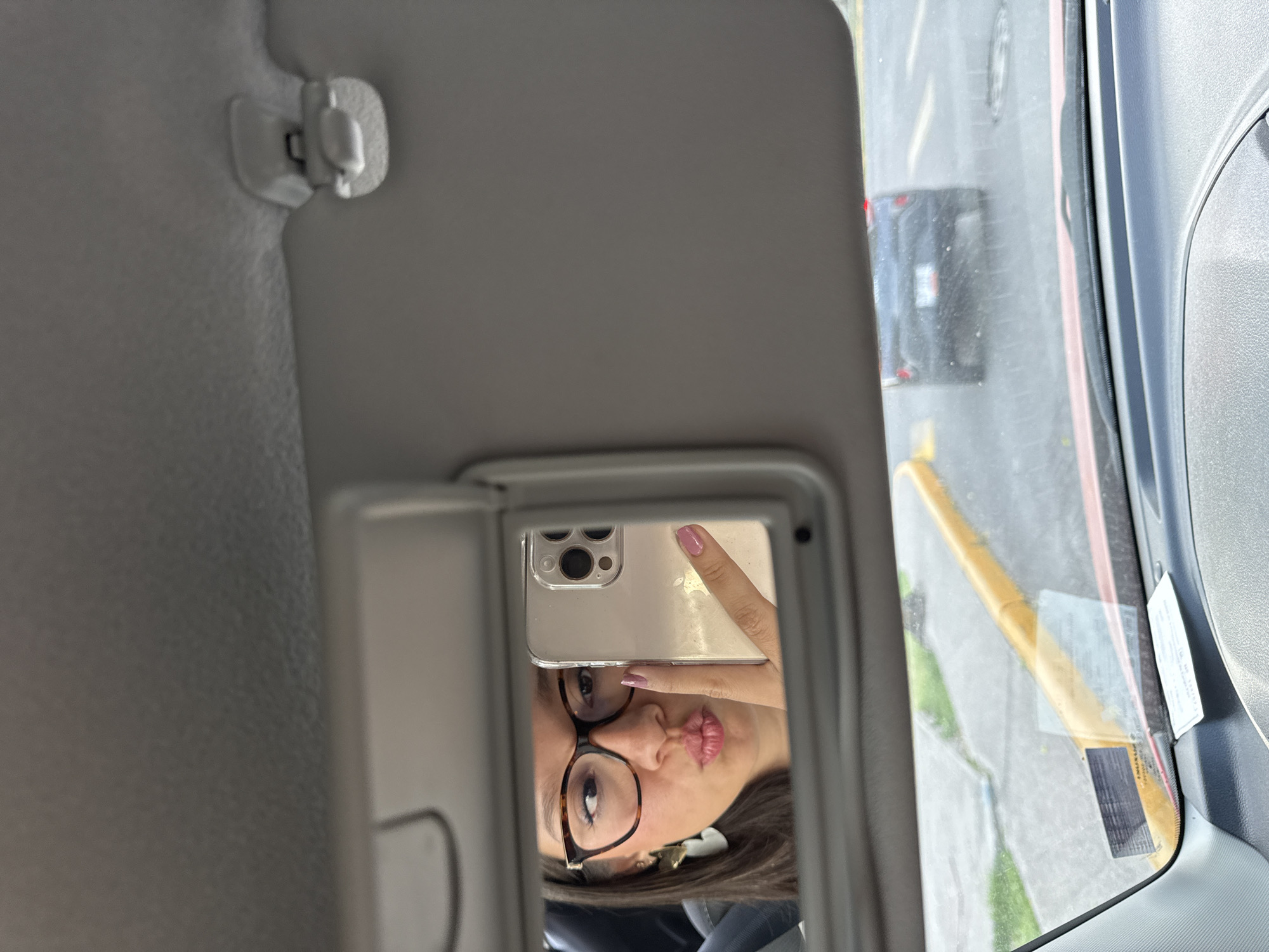 The image is taken from inside a car in San Bernardino Tlaxcalancingo, Mexico. The photo is a mirror selfie taken in the visor mirror. The reflection shows a person with glasses holding up a smartphone to take the picture. The person has dark hair and is making a kissing face. Outside the car window, there is a road with a yellow curb and a vehicle. The interior of the car is grey.