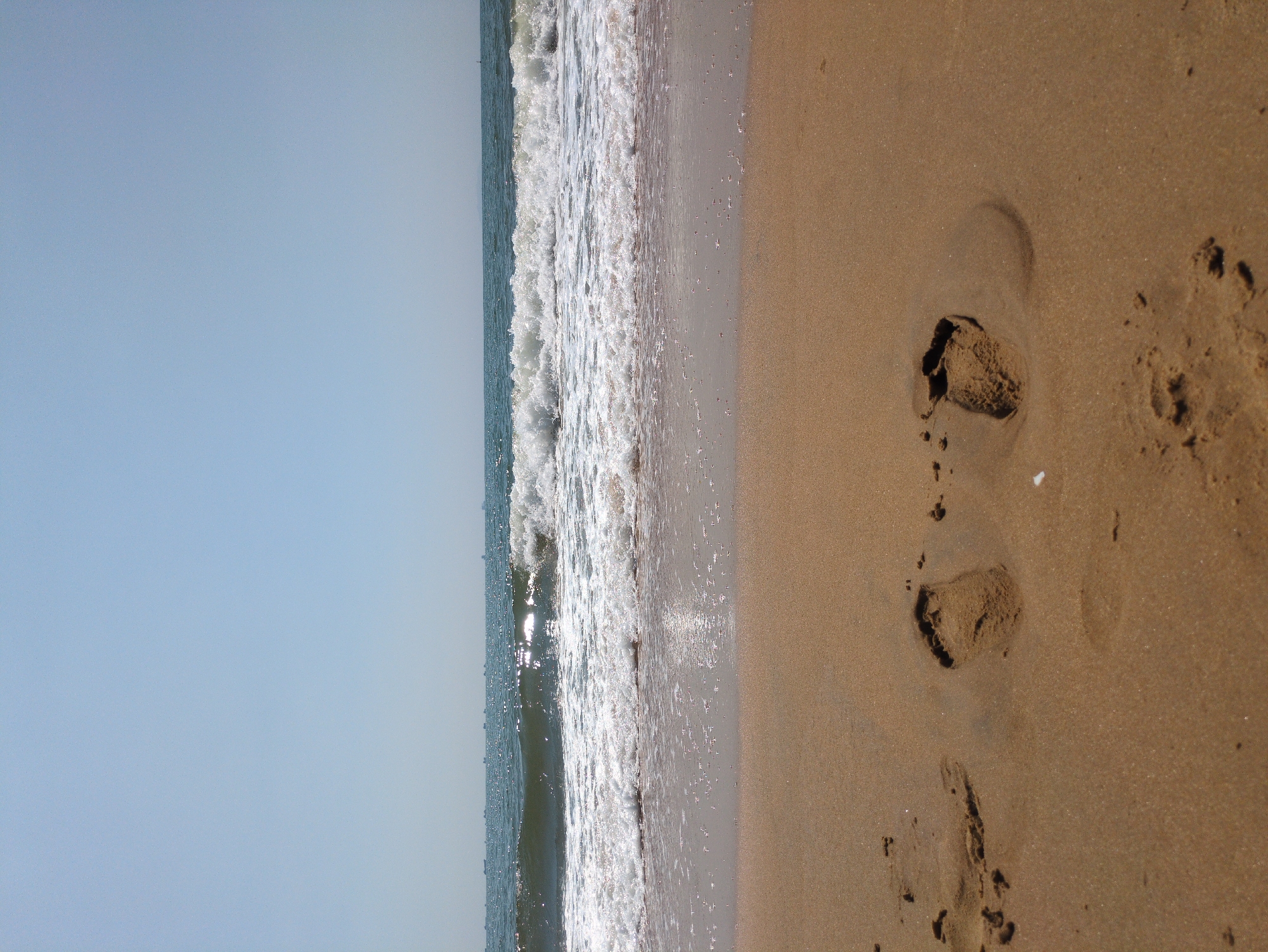 The image depicts a beach in South Quy Nhon Ward, Vietnam. The left side of the image shows a light blue sky. Adjacent to the sky is the ocean with waves. To the right is wet sand meeting the ocean, followed by drier sand with footprints.