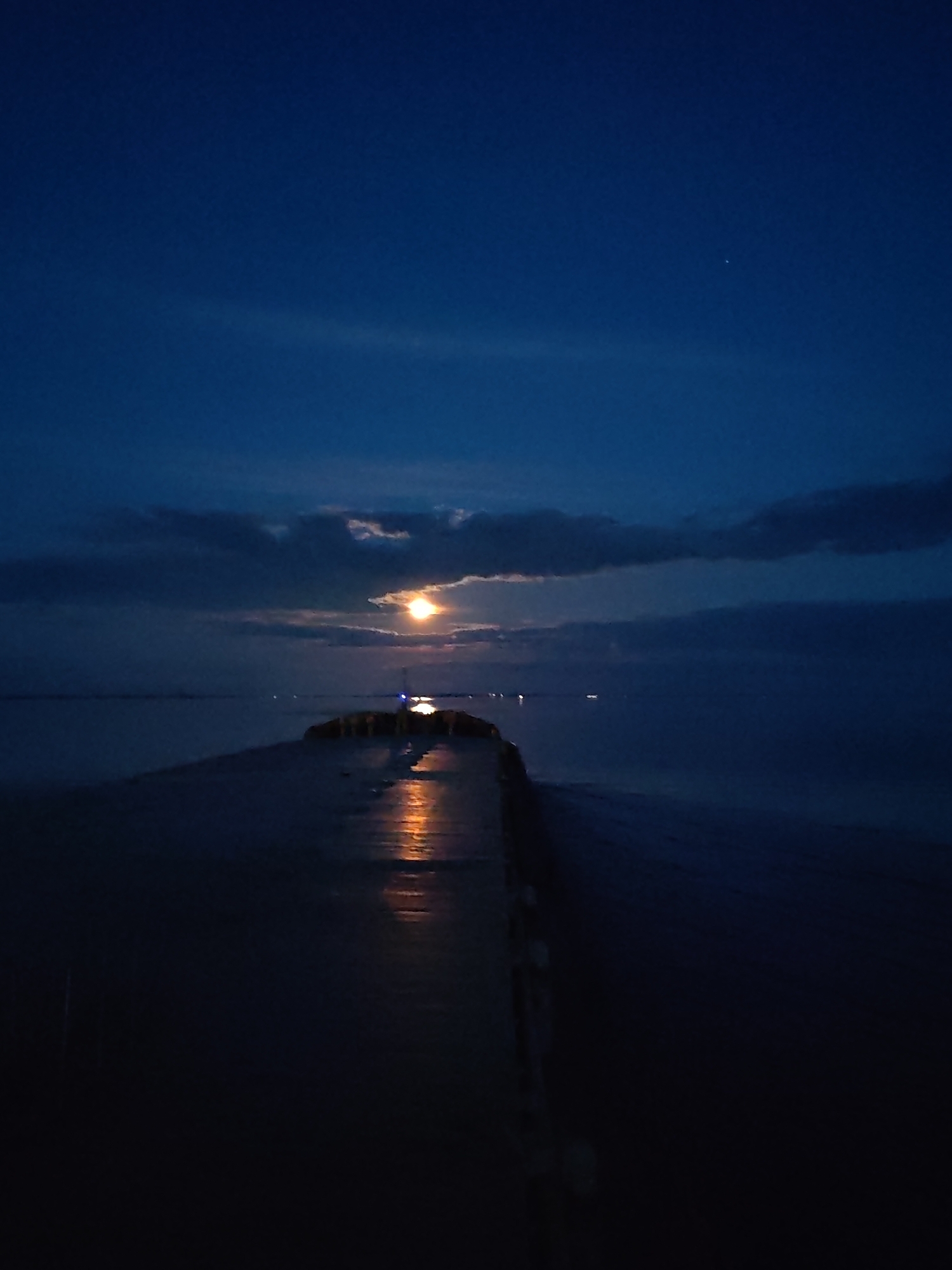 The image displays a serene nocturnal waterscape, likely a large body of water in Bol'shie Mkhi, Russia, under a moonlit sky. A prominent, glowing full moon is positioned in the upper mid-ground, partially obscured by dark, scattered clouds.
A long, dark structure, appearing to be a concrete pier or breakwater, extends from the foreground into the mid-distance, angling slightly to the right. Its wet surface brightly reflects the moonlight, creating a distinct orange-yellow streak that leads towards the moon. The water surrounding the structure is dark and calm, also reflecting some of the moon's light. In the distance, several small, indistinct lights are visible across the water, suggesting another shore or distant boats.
The sky is a deep, rich indigo blue, characteristic of a clear night, with darker clouds adding depth to the scene. There are no people or discernible text visible. The overall mood is tranquil, peaceful, and contemplative, highlighting the natural beauty of a moonlit night.