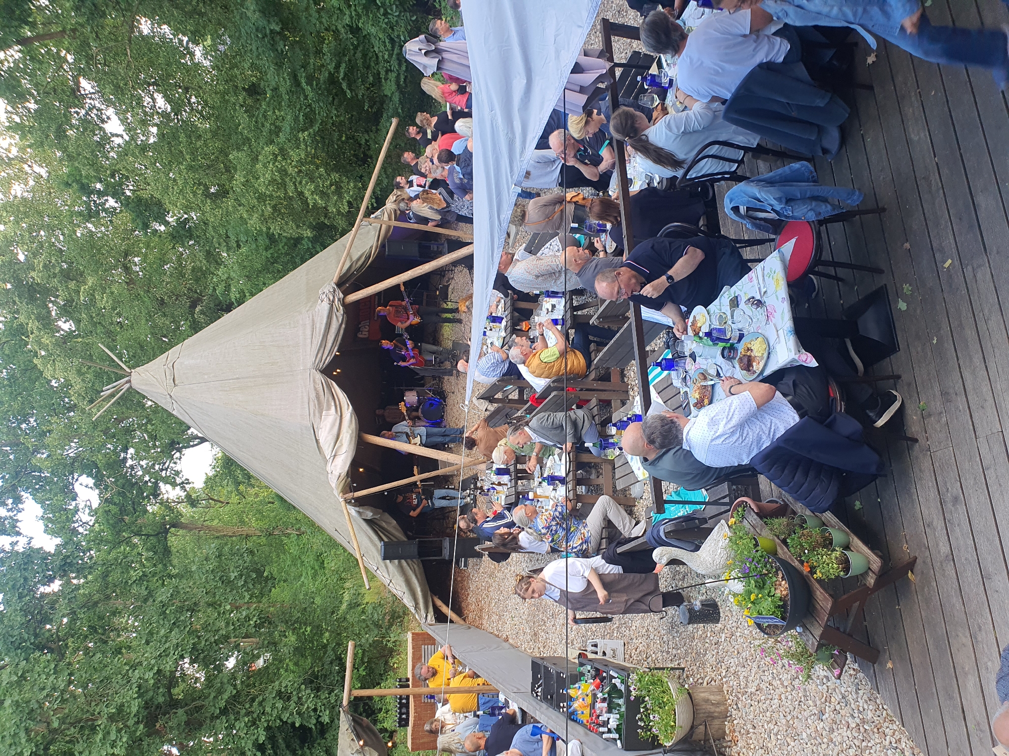 The image depicts an outdoor dining event at "Gartenwirtschaft am See" in Falkensee, Germany. A large, light-colored teepee-style tent is set up in the background, with a live band performing inside. The foreground and middle ground are filled with people seated at wooden tables, enjoying a meal and drinks. The tables are covered with patterned tablecloths and set with plates, glasses, and bottles. A woman in an apron is seen serving food, while other patrons are engaged in conversation and eating. The environment is rustic, with gravel ground and surrounding trees, suggesting a natural, park-like setting. The overall atmosphere appears to be relaxed and social, with a concert and dining experience taking place concurrently.