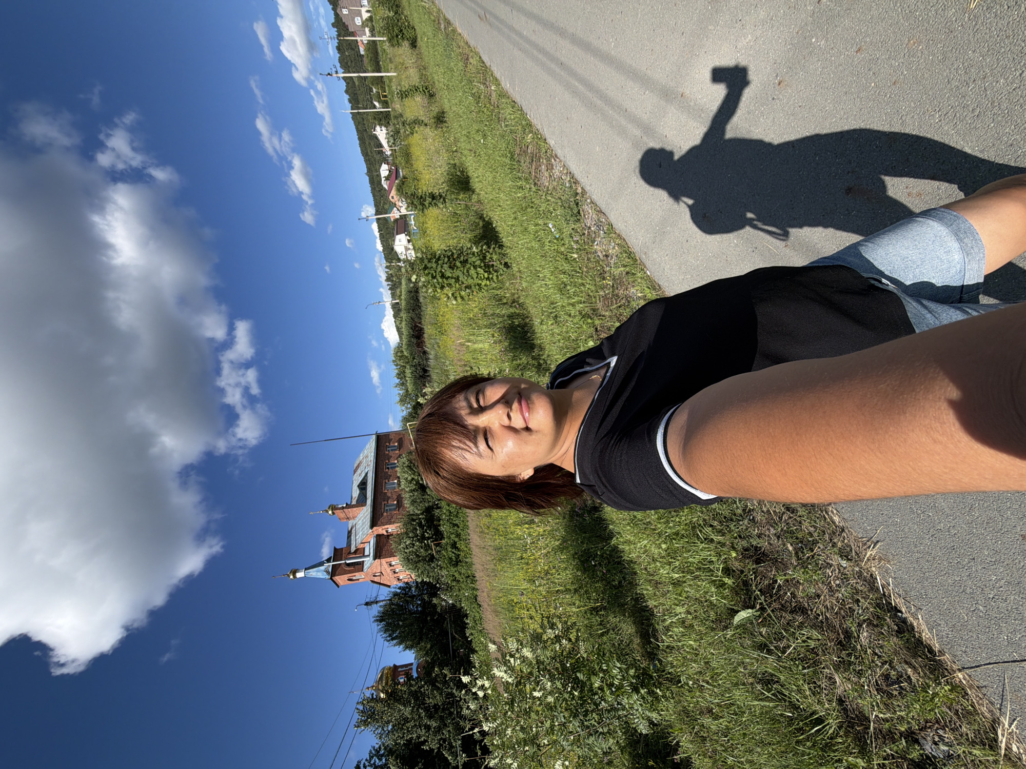 An outdoor, eye-level, full selfie taken by a woman with medium-length brown hair and a black v-neck t-shirt. She is wearing denim shorts and is standing on a paved road. To her left, there is a grassy hill with wild flowers and bushes. Further up the hill, a red brick church with a blue and gold spire can be seen. In the background, there are more houses and trees on a hillside, under a bright blue sky with fluffy white clouds. The woman's shadow is cast on the road to her right, holding a phone as if taking the selfie.