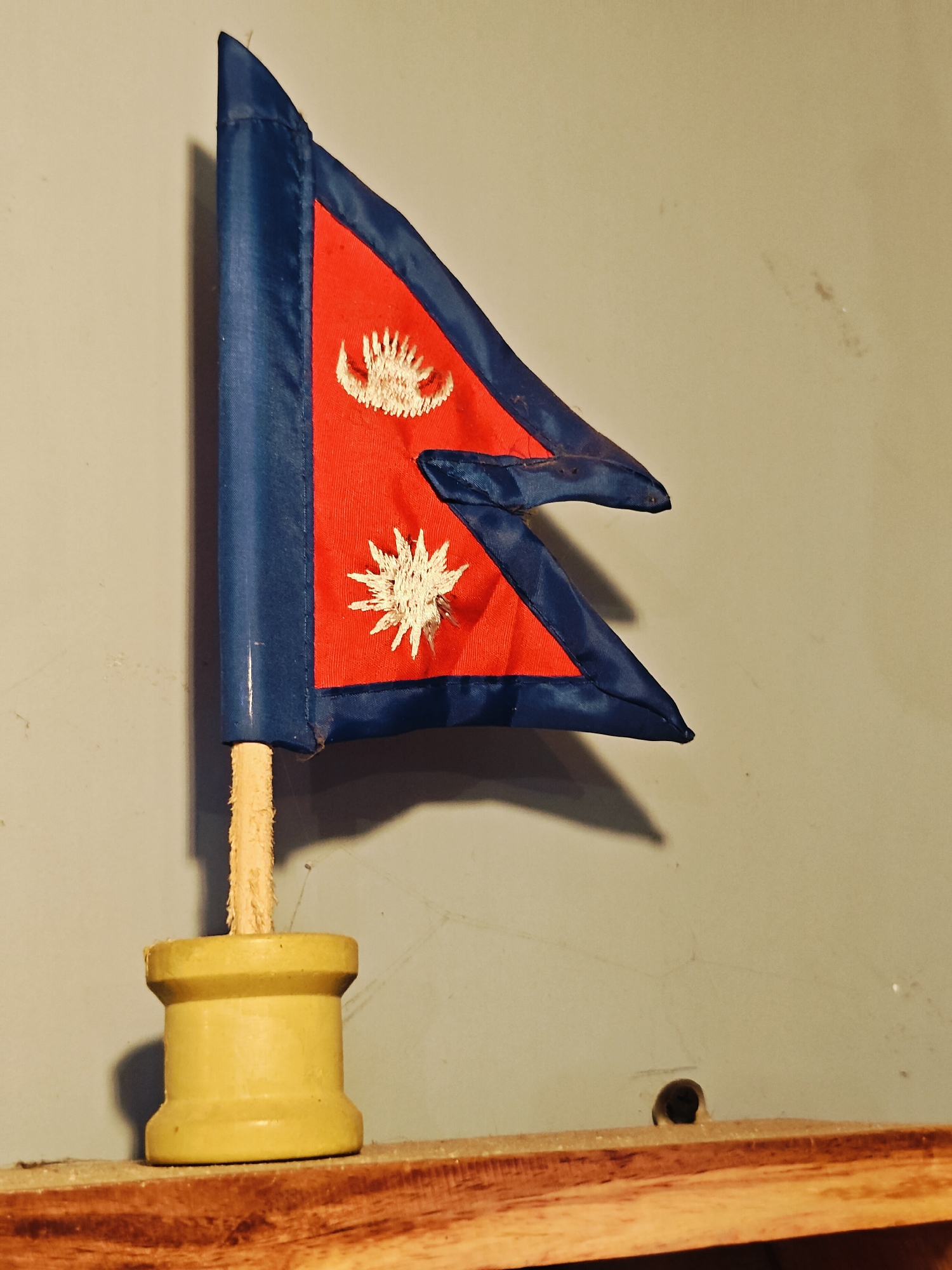 A small, decorative flag of Nepal is displayed on a wooden surface against a grey wall. The flag is crimson red with a blue border and features embroidered white symbols of the sun and moon. It is attached to a wooden stick that is inserted into a yellow, cylindrical base. The wall behind the flag shows some texture and faint smudges, and a small, dark hole is visible in the lower right portion of the wall. The overall scene suggests an indoor display. This image is not located in Lalitpur, Nepal.