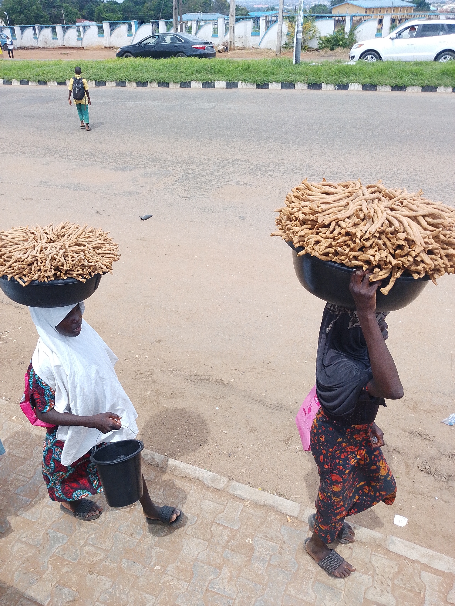 In Oja Oba, Oko Erin, Nigeria, two women are seen walking while carrying large bowls of food on their heads. The woman on the left wears a white hijab, a colorful patterned dress, and carries a black bucket in her hand. The woman on the right wears a black head covering, a patterned skirt, and has a pink bag slung over her shoulder. Both women are wearing sandals. They are walking on a paved sidewalk next to a dusty road. In the background, there is a street with cars, including a black sedan and a white SUV. Buildings and trees can be seen in the distance. The scene appears to be set during the day, under natural light, suggesting fair weather. The overall impression is that of a busy marketplace or roadside scene in Nigeria, with vendors engaged in their daily trade.
