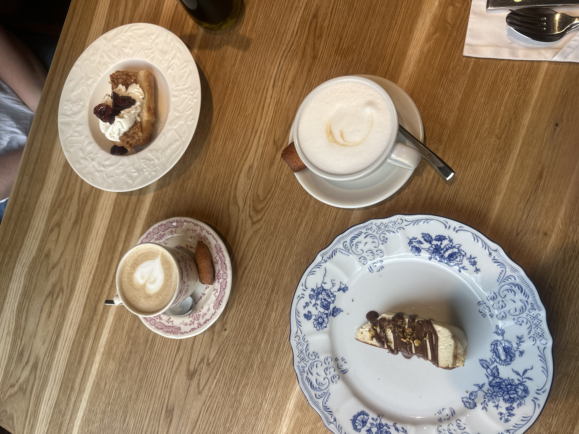 A high-angle, close-up shot shows two cups of coffee and two plates of dessert on a wooden table. The coffee cups are white with saucers. One saucer is white with a pink floral pattern, and the other is plain white. Both cups contain latte art; one is a heart shape, and the other is a swirl. A small, oblong cookie rests on each saucer.
One dessert is a slice of cake or pastry topped with whipped cream and what appears to be dried berries. This is on a white plate with an embossed pattern. The other dessert is a slice of cake with a cream filling, drizzled with chocolate sauce and sprinkled with nuts. This is on a white plate with a blue floral pattern. Cutlery, including a fork and spoon, are visible on a white napkin in the lower right corner of the image.
The setting is likely a cafe or restaurant. Given the location provided, this scene could be from a cafe in Petrozavodsk, Russia. The lighting suggests an indoor environment, possibly with natural light from a nearby window.