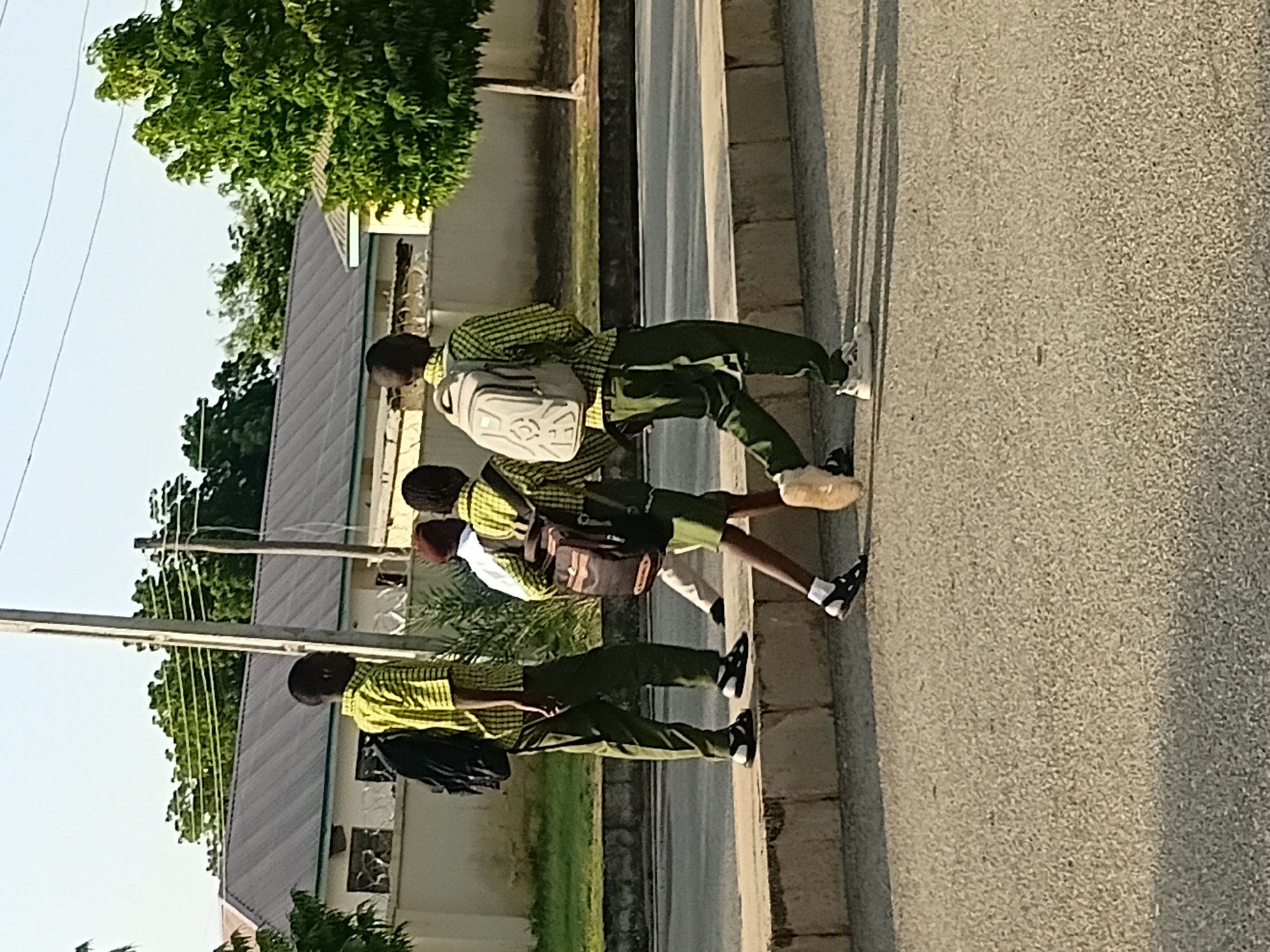 Three young male students, appearing to be in their early to mid-teens, are walking on a paved sidewalk next to a road. They are dressed in school uniforms consisting of yellow and green checkered shirts and green pants. Two of the students are carrying backpacks. The student on the left has a black backpack, the student in the middle has a dark brown bag slung over his shoulder, and the student on the right has a light gray backpack. They are walking from left to right in the image, and their shadows are cast on the road. In the background, there is a building with a corrugated metal roof, some trees, and a utility pole. The scene appears to be in an urban or semi-urban setting, consistent with Jalingo, Nigeria, based on the architecture and vegetation. The time of day appears to be late afternoon, indicated by the long shadows.