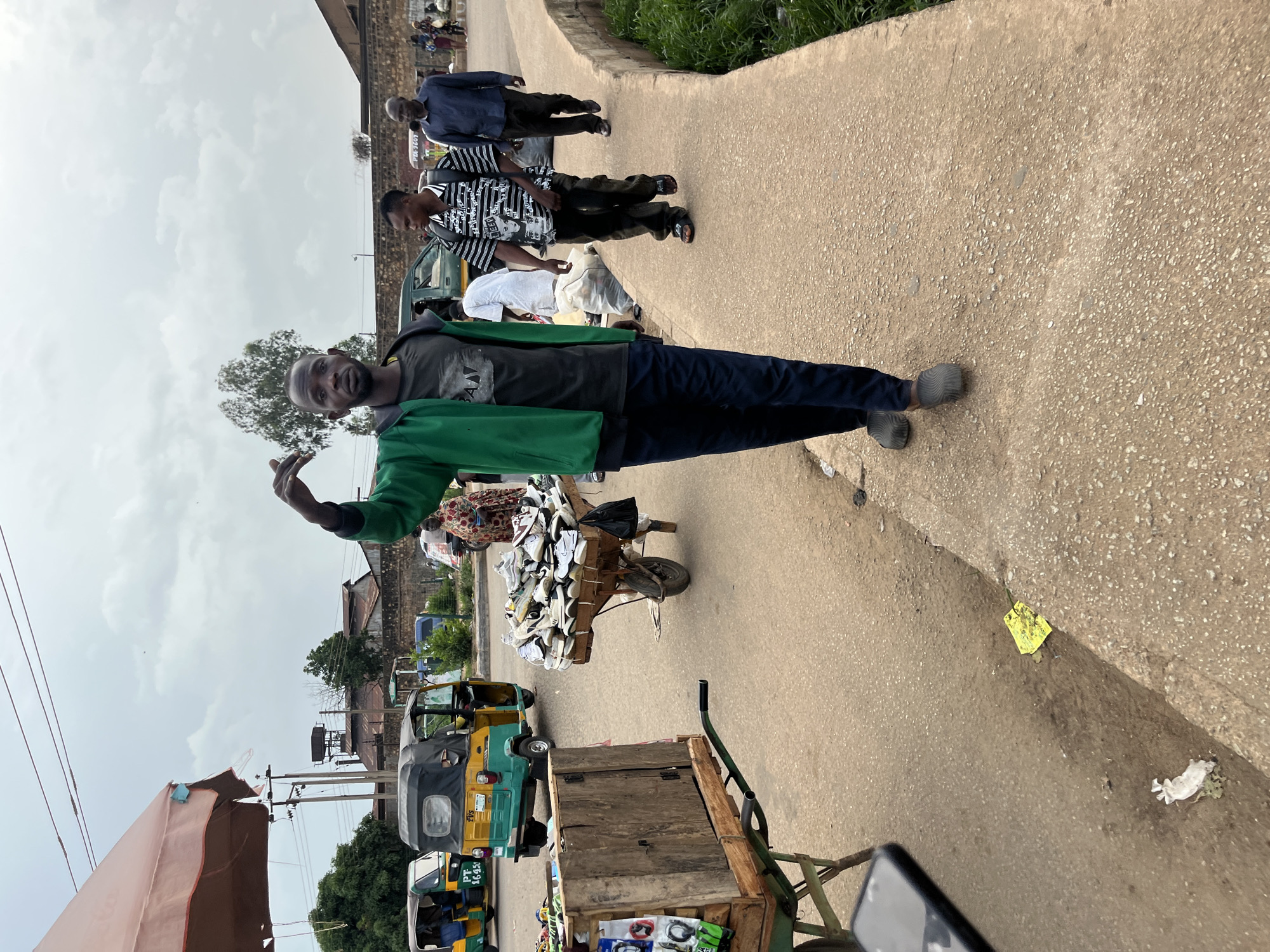 A busy street scene in Bukuru, Nigeria, depicts a young man in a green jacket and dark pants walking towards the camera, gesturing with his right hand. He is the central figure in the image. To his left, a cart is piled high with shoes, and another cart is laden with a wooden structure. In the background, several auto-rickshaws are visible, along with other pedestrians and buildings. The sky is overcast, suggesting a cloudy day. The ground is paved, with some debris and grass on the side. The overall impression is one of a lively marketplace or transit hub.