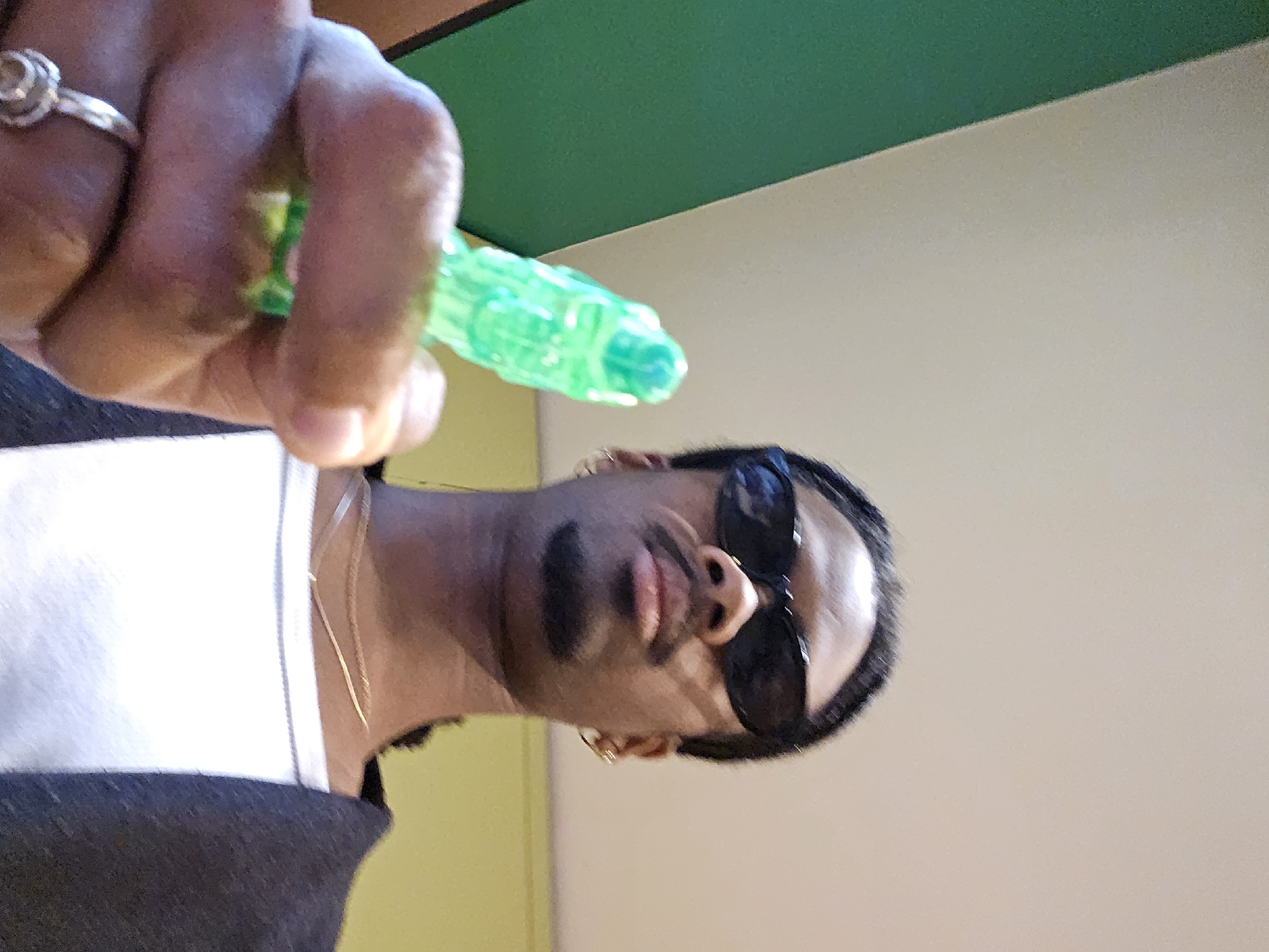 A person with dark skin, wearing dark sunglasses, a white and black shirt, and gold jewelry is holding a green plastic object in their hand. The background consists of a green and beige wall. The location of the image cannot be determined as the image lacks the necessary environmental context to establish its whereabouts in Paris, France.