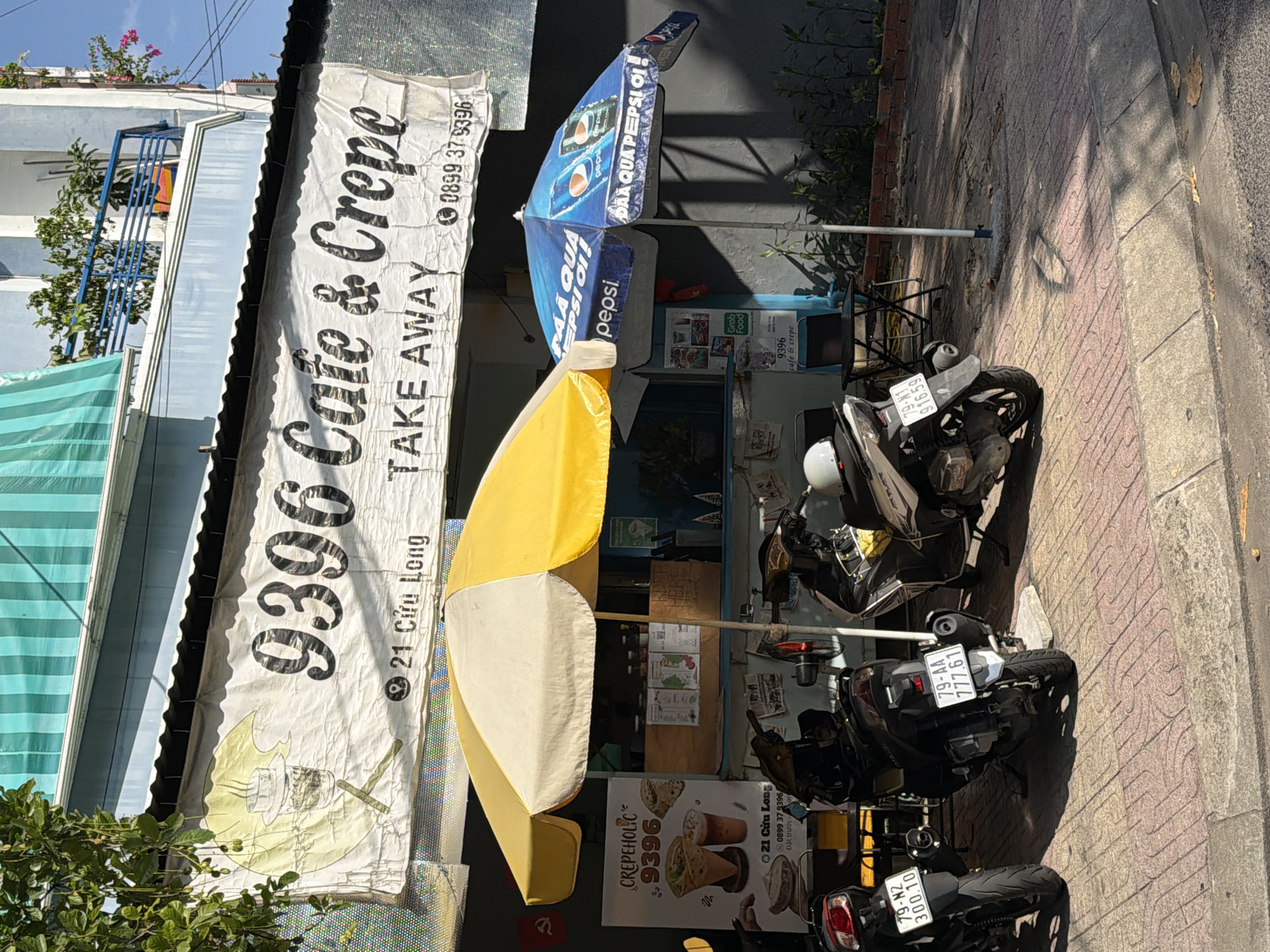 This image shows a storefront in Nha Trang, Vietnam, with a banner reading "9396 Cafe & Crepe TAKE AWAY." The cafe appears to be a small, open-air establishment with a counter and seating area. Two motorcycles are parked in front of the cafe, and a yellow and white umbrella provides shade over part of the seating area. A blue Pepsi umbrella is also visible. A sign on the counter advertises "CREPEHOLIC 9396." The street is paved with red and pink tiles. The building in the background has a light blue facade and a balcony with plants. The sky is blue and sunny.