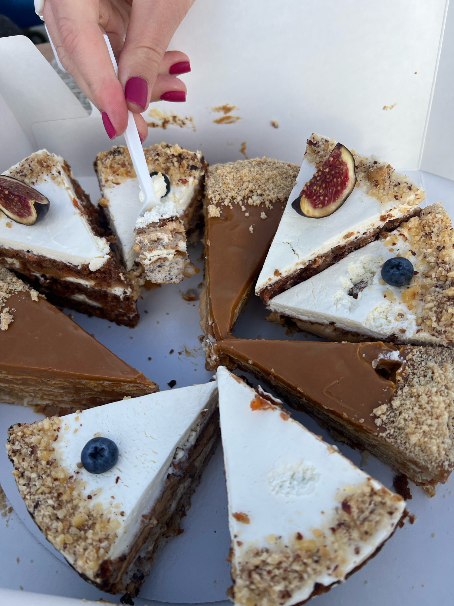 In Saint Petersburg, Russia, a close-up shot captures a hand with pink nail polish using a white plastic fork to take a bite from a slice of cake. The cake is layered with white frosting and what appears to be caramel or dulce de leche, topped with chopped nuts and a blueberry. Other slices of cake are arranged in a white box, some garnished with a slice of fig and another blueberry. The background is out of focus, showing a white surface.
