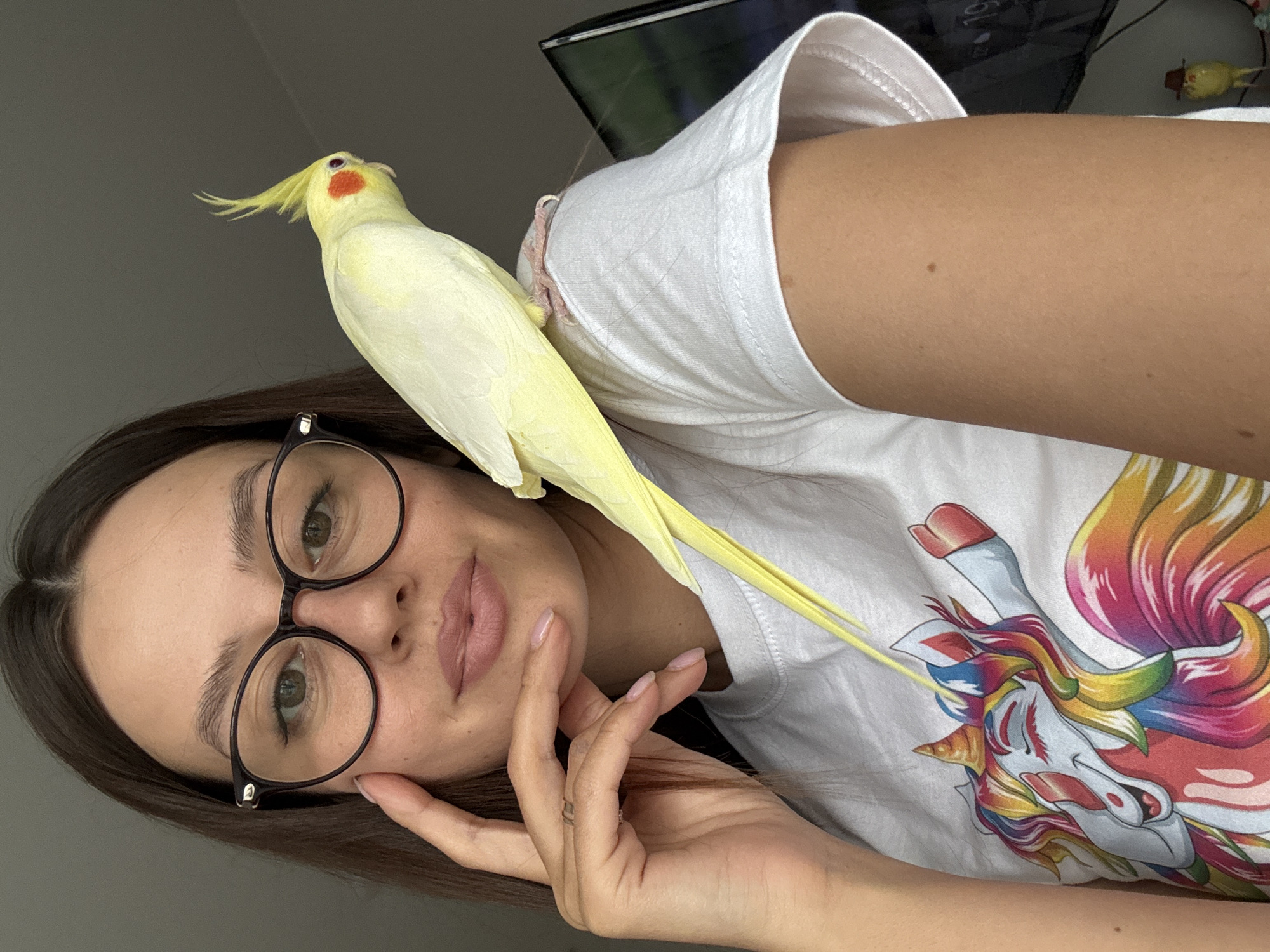 A young woman with dark brown hair and glasses is pictured in a close-up, over-the-shoulder shot. She is wearing a white t-shirt with a colorful unicorn graphic. A light yellow cockatiel is perched on her left shoulder, with its tail feathers extending down her back. The woman has her right hand near her face, with her fingers touching her cheek. The background appears to be an indoor setting with neutral-colored walls and a television screen visible on the right side of the frame. The image is taken from a slightly elevated angle. The scene does not provide any specific indicators of its location in Krasnoyarsk, Russia.