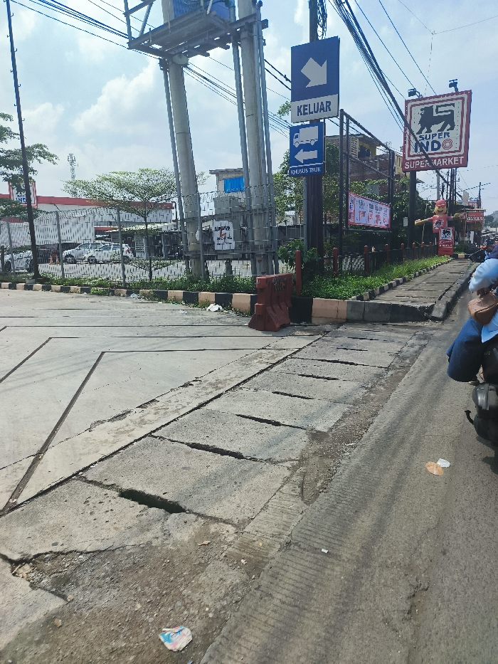 The image shows a street in Ngamprah, Indonesia, at the coordinates -6.868 latitude and 107.524 longitude. The street is paved with gray concrete and has a series of drainage channels running down the middle. There is a signpost at the street corner with a blue sign that reads "KELUAR" in white letters and a symbol of a truck with an arrow pointing left below. The sign is attached to a metal pole with electrical wires running overhead. On the right side of the street, there is a sidewalk with a sign for "SUPER INDO SUPER MARKET" in yellow and black. A person in blue clothing is standing on the sidewalk, facing away from the camera. Their right hand is resting on the handlebars of a motorcycle. The motorcycle is parked on the sidewalk, facing away from the camera. There is a concrete barrier on the left side of the street. There is a red concrete block behind the barrier. The image is taken from a slightly elevated angle, looking down the street. The sky is blue with some clouds. The trees are green. The day is clear and sunny. The pavement is gray and slightly cracked. There is a small piece of trash on the ground.