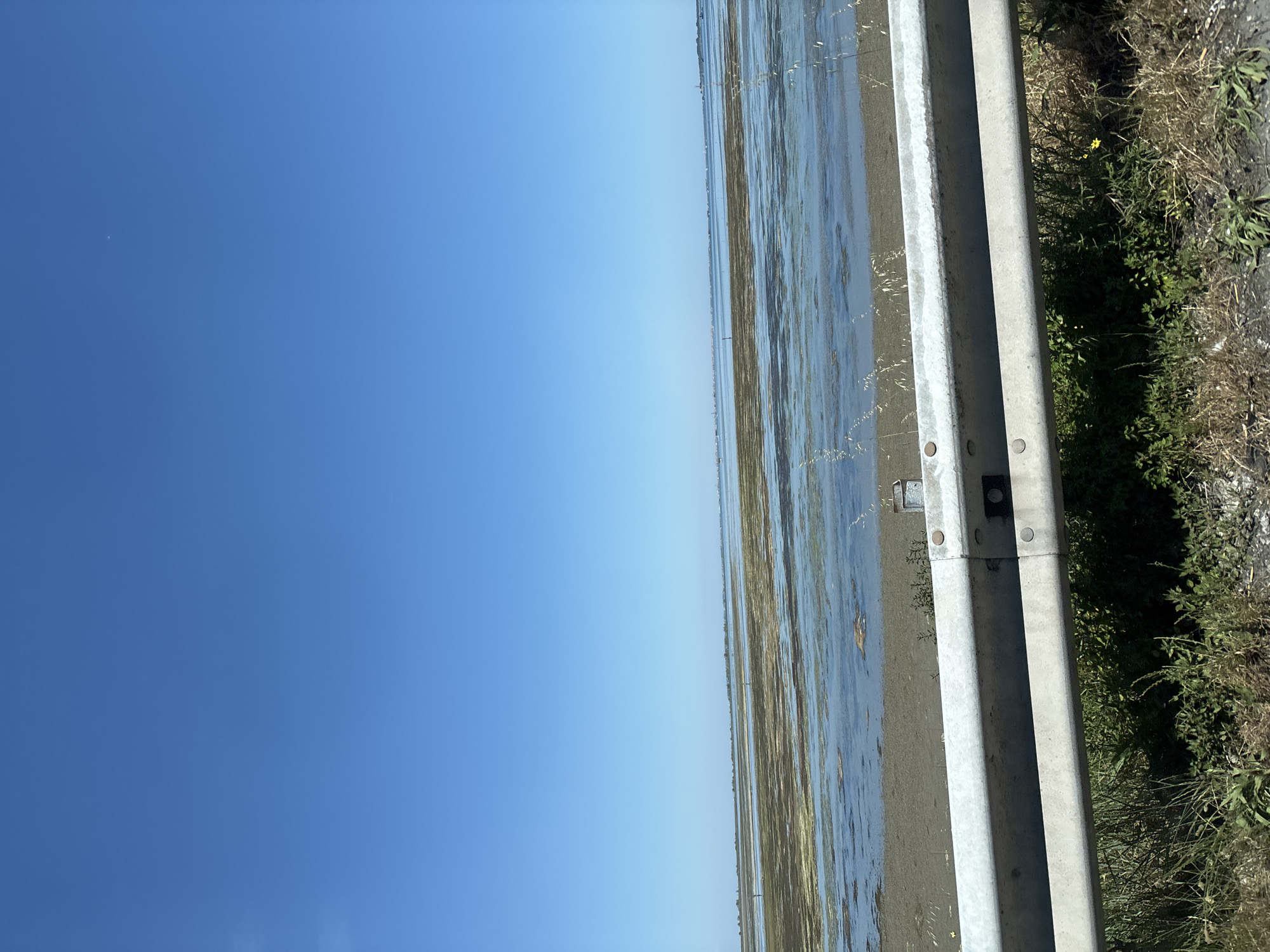 A photograph taken in Chioggia, Italy, presents a landscape divided between the sky and an adjacent field. The sky is a uniform, bright blue and occupies more than half of the left side of the image. A gray, weathered guardrail is situated vertically, separating the sky from the field. The field appears flooded, with the water level high enough to reflect the sky and the surrounding area, giving it a mirrored effect. Vegetation lines the edge of the field. The photograph captures a rural or semi-rural setting where agricultural land meets the sky, bordered by infrastructure.
