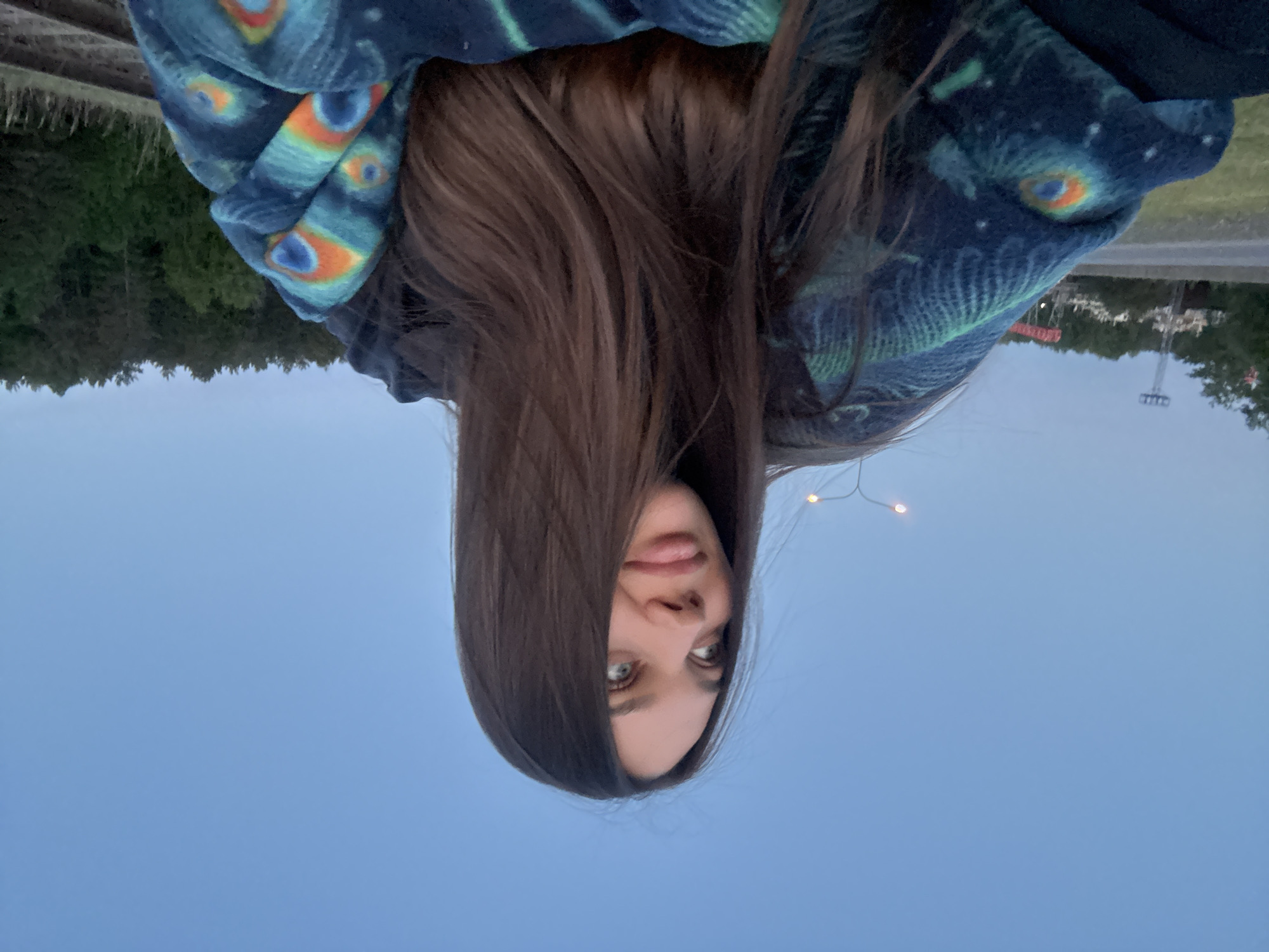 A selfie of a young woman with long brown hair, wearing a blue and turquoise patterned jacket or hoodie. She is outdoors, likely in Chaikovsky, Russia, based on landscape features in the background. The picture is oriented upside down. The woman has light eyes and fair skin. She is looking directly at the camera. The background shows trees, a road, some buildings, and a light pole. The sky is a pale blue.