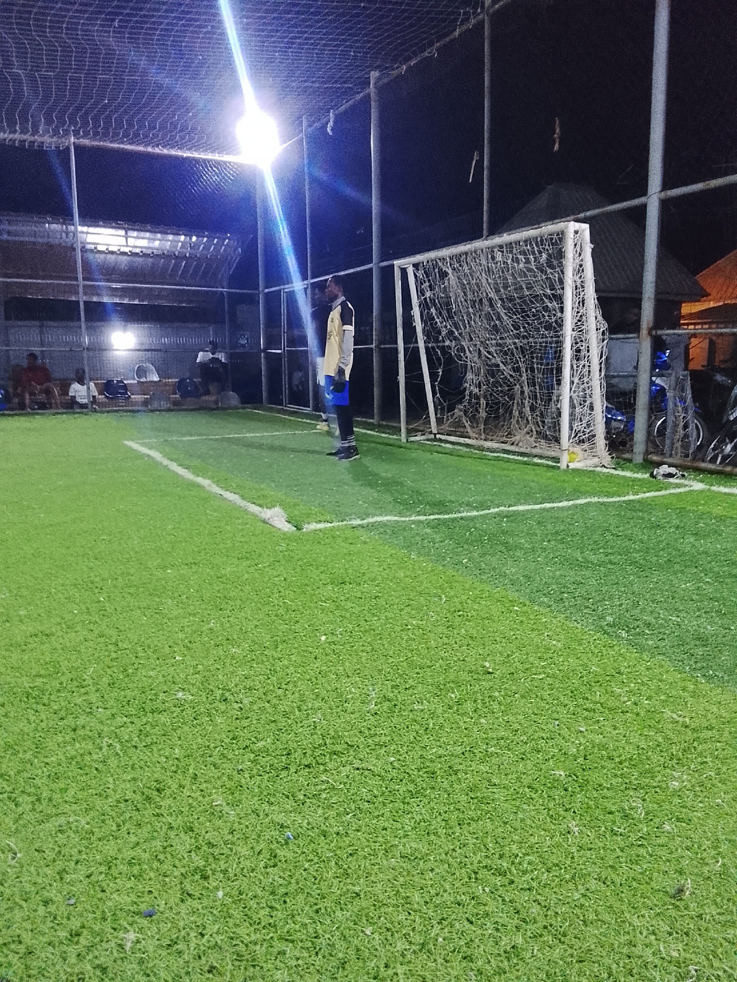 The image depicts an artificial turf soccer field at night, likely in Akwanga, Nigeria. A man in a light-colored jersey and blue shorts stands near a goal, facing away from the camera. The field is enclosed by a wire mesh fence. A goal with a net stands to the right of the man. Behind the fence and goal are motorbikes. A group of people are seated on a bench under a covered area in the background. Overhead lighting illuminates the field.