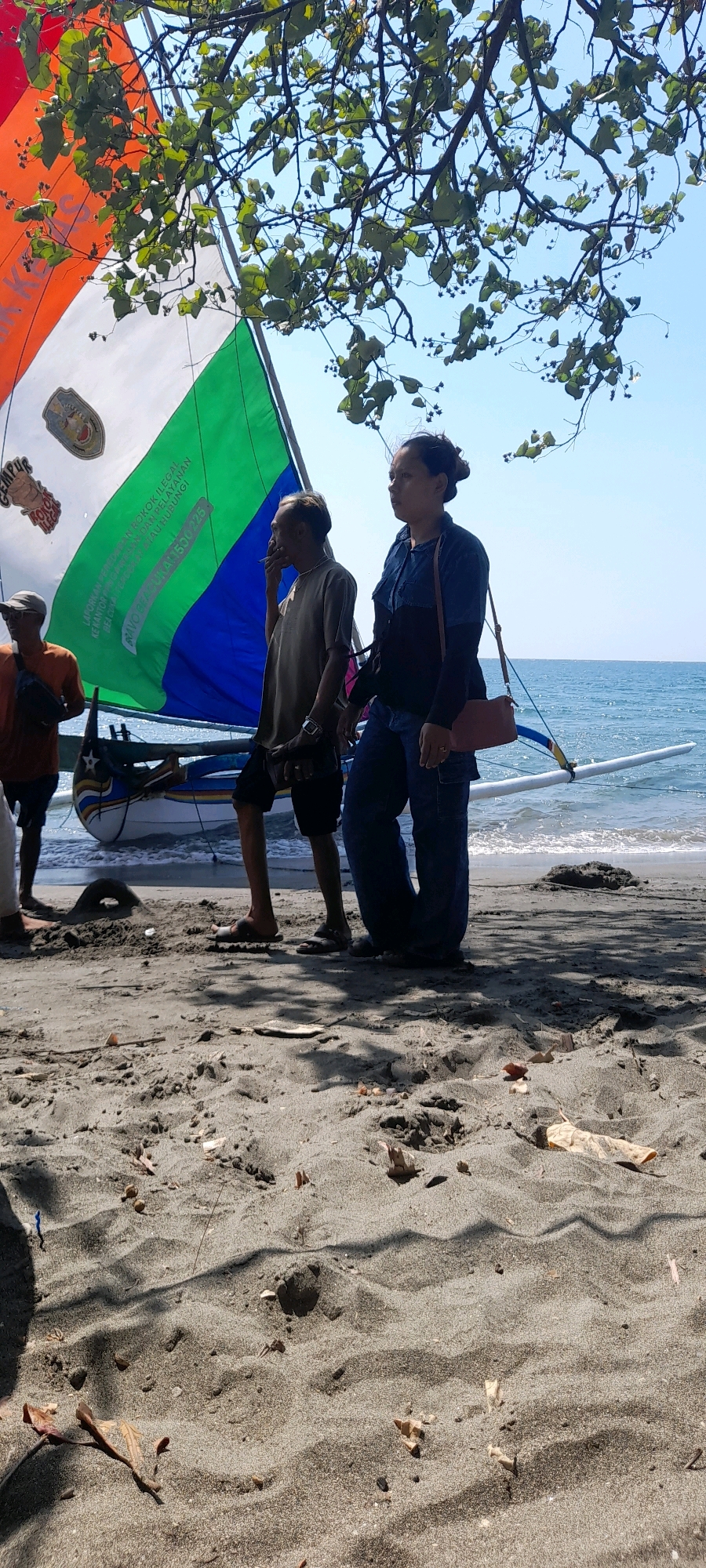 Two individuals, a male and a female, stand on a sandy beach next to a colorful sailboat. The male, positioned slightly in front and to the left of the female, is wearing a tan t-shirt and dark shorts, holding a cigarette in his right hand. He has a watch on his left wrist. The female, to his right, is wearing a dark blue collared shirt with long sleeves and dark pants. A small brown purse is slung over her right shoulder. Their gaze is directed towards the right of the frame. The sailboat behind them has a large sail divided into orange, white, green, and blue sections, with text and a crest visible on the white portion. The boat rests on the sand near the water's edge. The beach sand is dark and textured, with some scattered leaves and debris. In the background, the blue ocean stretches to the horizon under a clear sky, with a tree branch casting shadows on the sand in the foreground. The scene appears to be set at Pasir Putih beach in Indonesia.